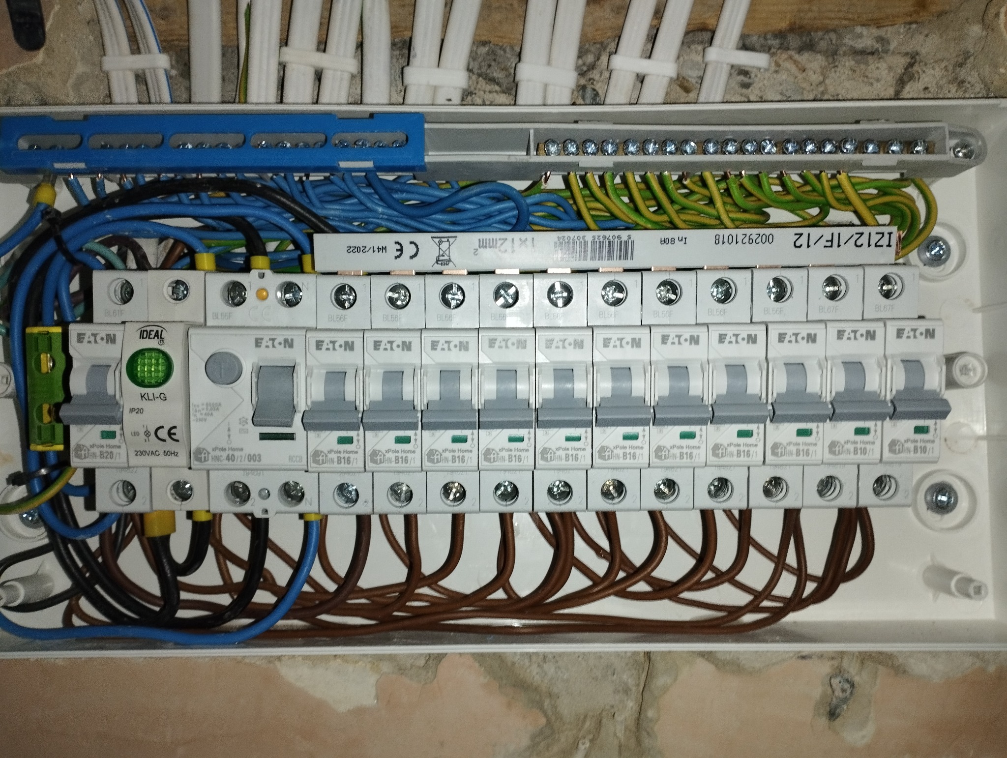 Rozdzielnica elektryczna z zainstalowanymi bezpiecznikami i podłączonymi przewodami w kolorach niebieskim, brązowym, zielono-żółtym i białym, widoczne listwy zaciskowe i oznaczenia producentów.