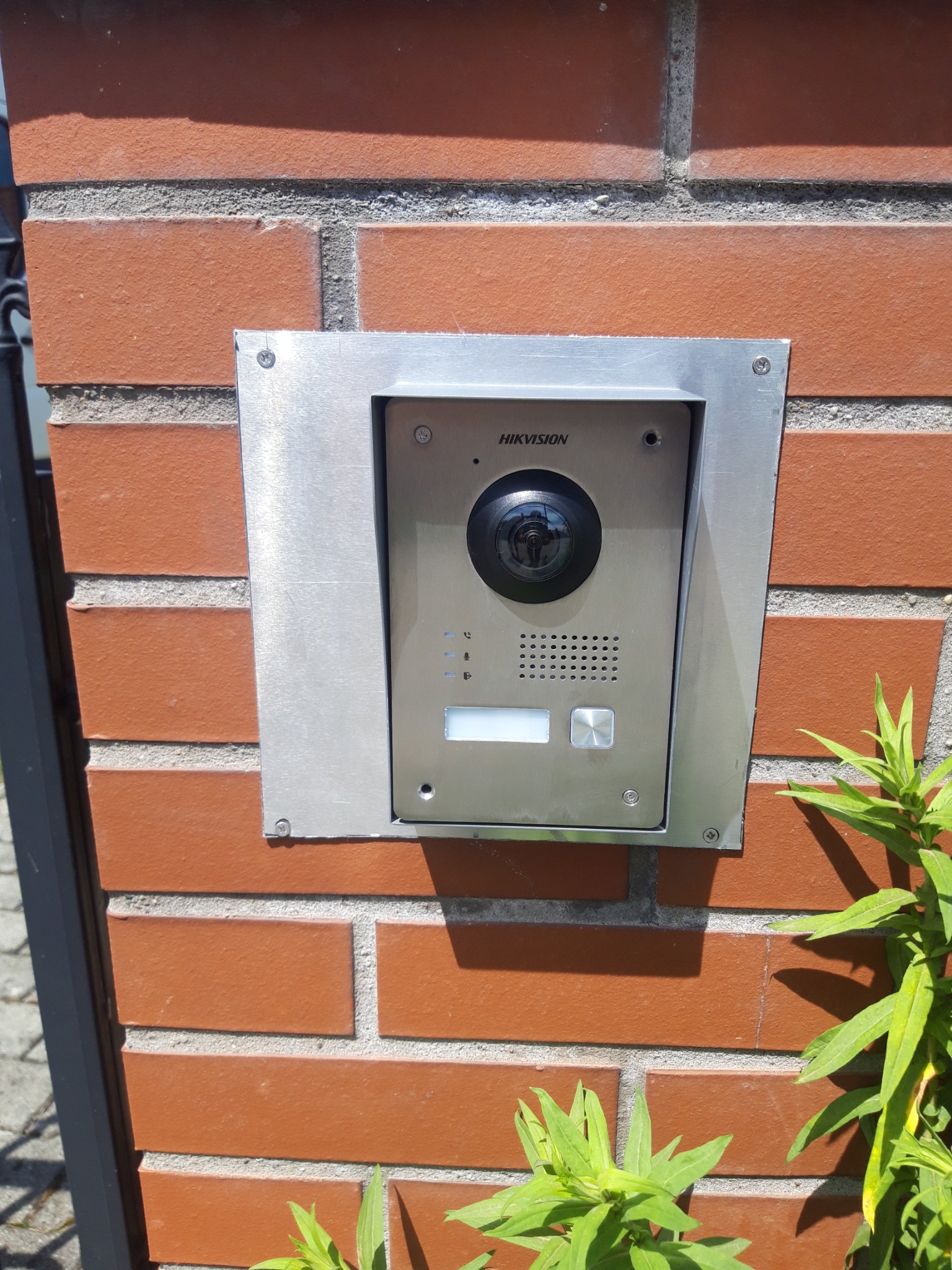 Zewnętrzny panel wideodomofonu Hikvision zamontowany na ceglanym słupku ogrodzenia, widoczne śruby mocujące i fragment rośliny
