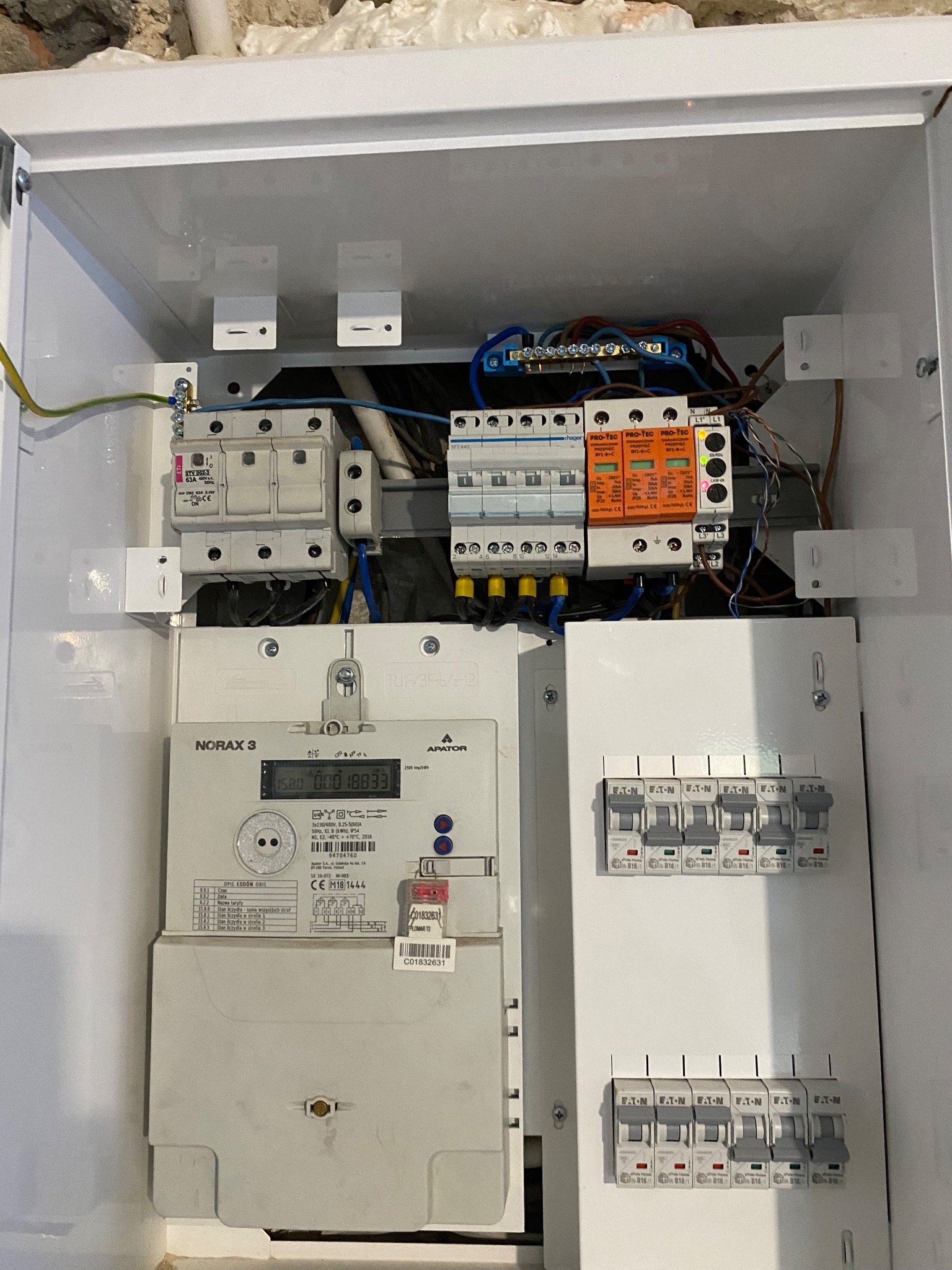 Zamknięta skrzynka elektryczna z licznikiem Norax 3, zabezpieczeniami Pro-Tec i wyłącznikami Eaton. Widoczne przewody i elementy instalacji elektrycznej.