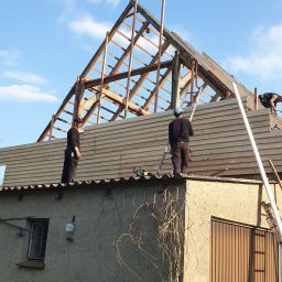 Dach i naprawa - Ekipa dekarzy montuje drewnianą boazerię na dachu małego budynku z widoczną starą więźbą dachową i częściowo ułożonym pokryciem.