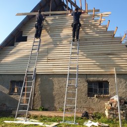 Dach i naprawa - Dwóch pracowników na drabinach montuje drewnianą konstrukcję dachu na budynku z oknami, w tle błękitne niebo.