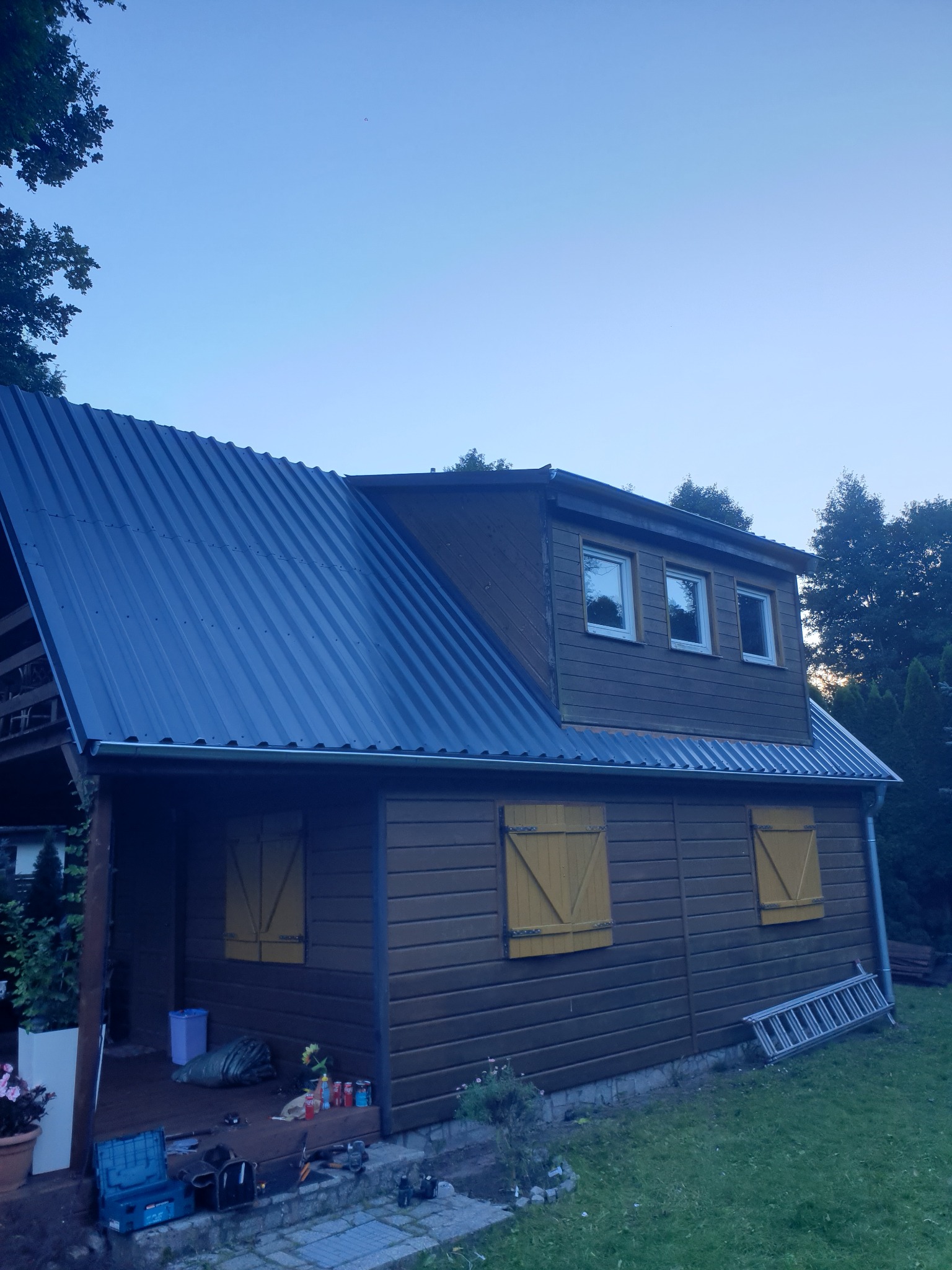 Dom z nowym, błyszczącym, ciemnoszarym dachem z blachy trapezowej, żółte okiennice, widoczne narzędzia i drabina, częściowo zachmurzone niebo.