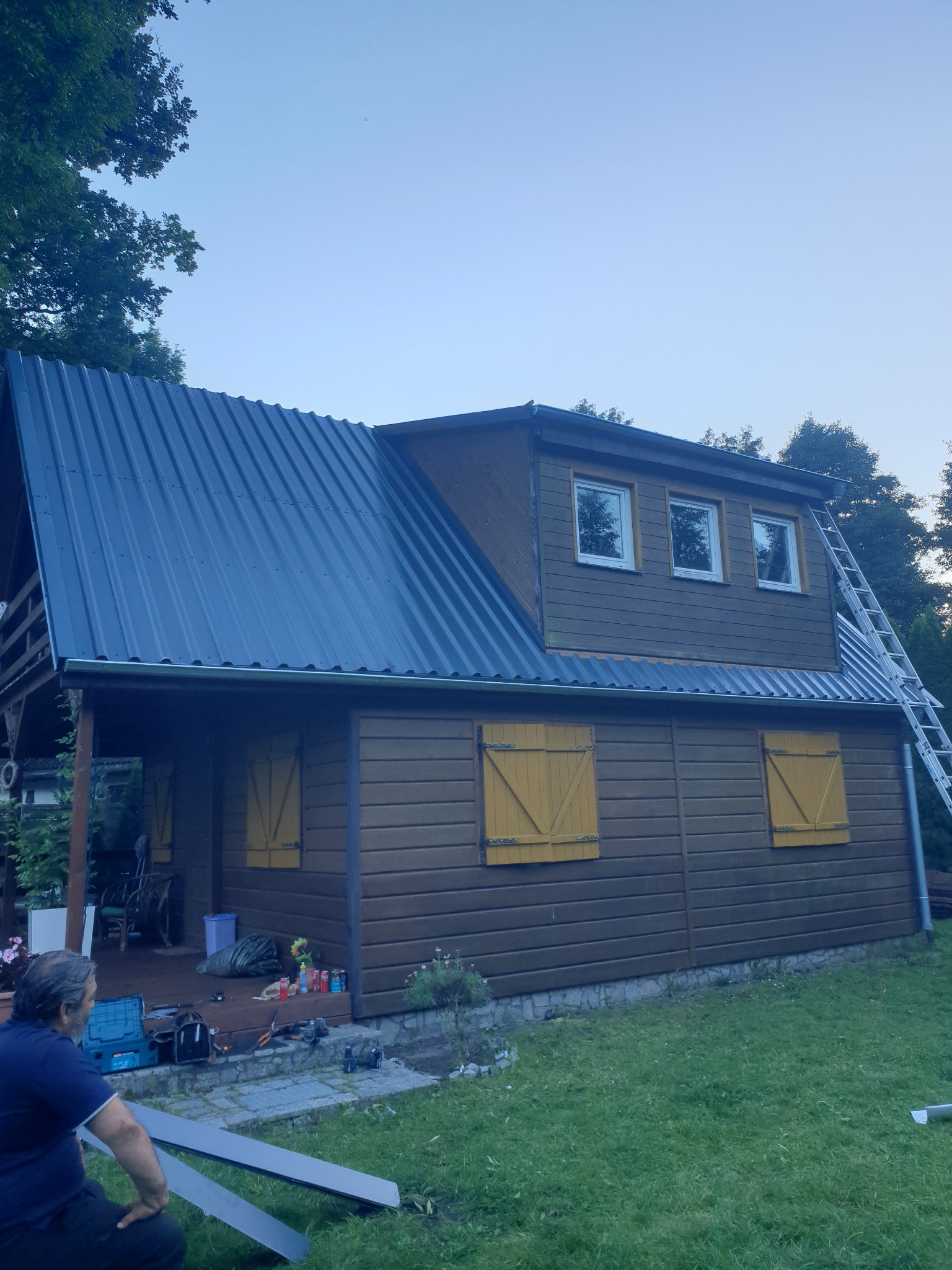 Dom z nowym, ciemnym dachem z blachy trapezowej i zamontowanymi rynnami. Na dachu drabina, a na ziemi narzędzia i materiały przygotowane do pracy. Widoczny fragment osoby pracującej przy dachu.