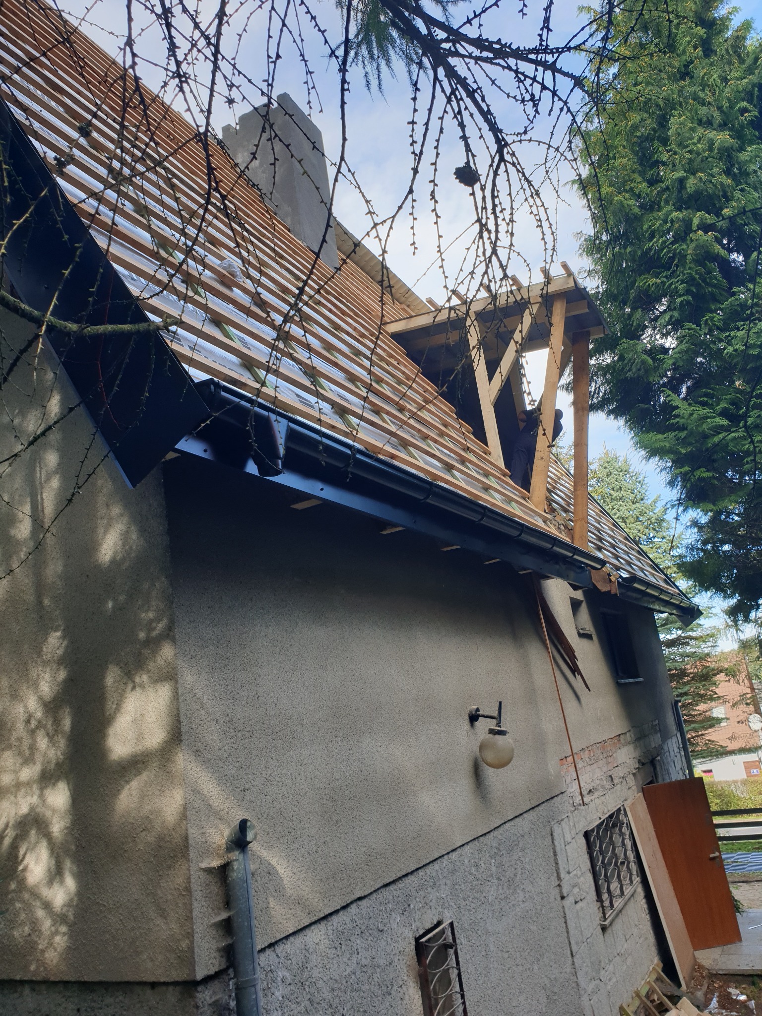 Budynek z widoczną konstrukcją dachu w trakcie budowy, z elementami drewnianymi i membraną, z zamontowaną rynną w kolorze grafitowym, na tle drzew i błękitnego nieba.