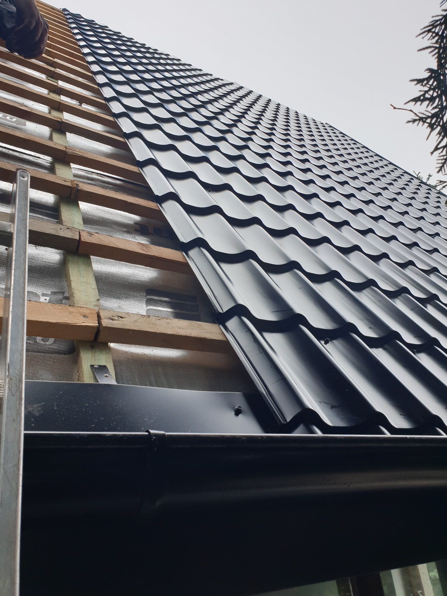 Montaż czarnej blachodachówki na drewnianej konstrukcji dachu z widoczną membraną, fragment rynny w dolnej części kadru.