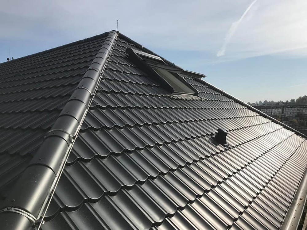 Czarny dach z dachówki ceramicznej z oknami dachowymi, widok z góry, w tle błękitne niebo z lekką smugą kondensacyjną.