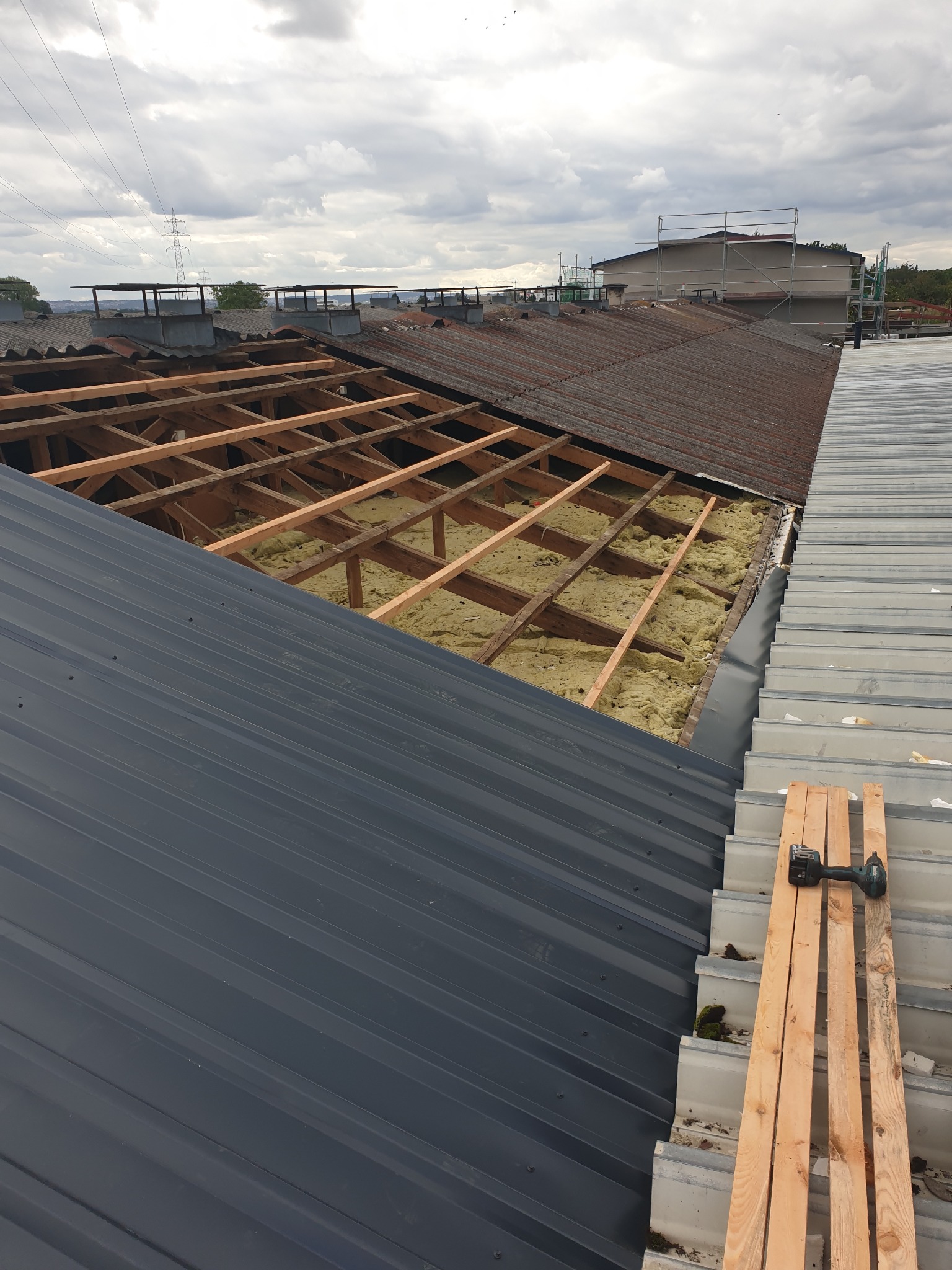 Częściowy widok dachu w trakcie prac: nowa blacha trapezowa, odsłonięta więźba z izolacją, fragment starego dachu z kominami w tle, narzędzie na deskach.