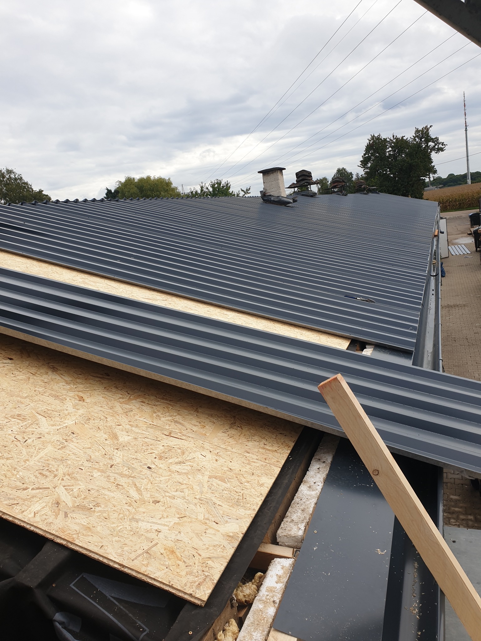 Montaż płyt OSB na dachu z blachy trapezowej, widoczny fragment komina i krajobraz w tle.