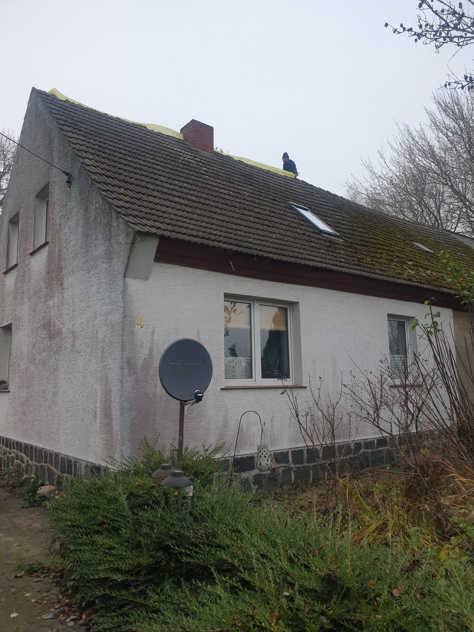 Dom jednorodzinny z częściowo zdjętym starym pokryciem dachowym, widoczna żółta membrana dachowa i pracownik na dachu. Część dachu pokryta mchem.