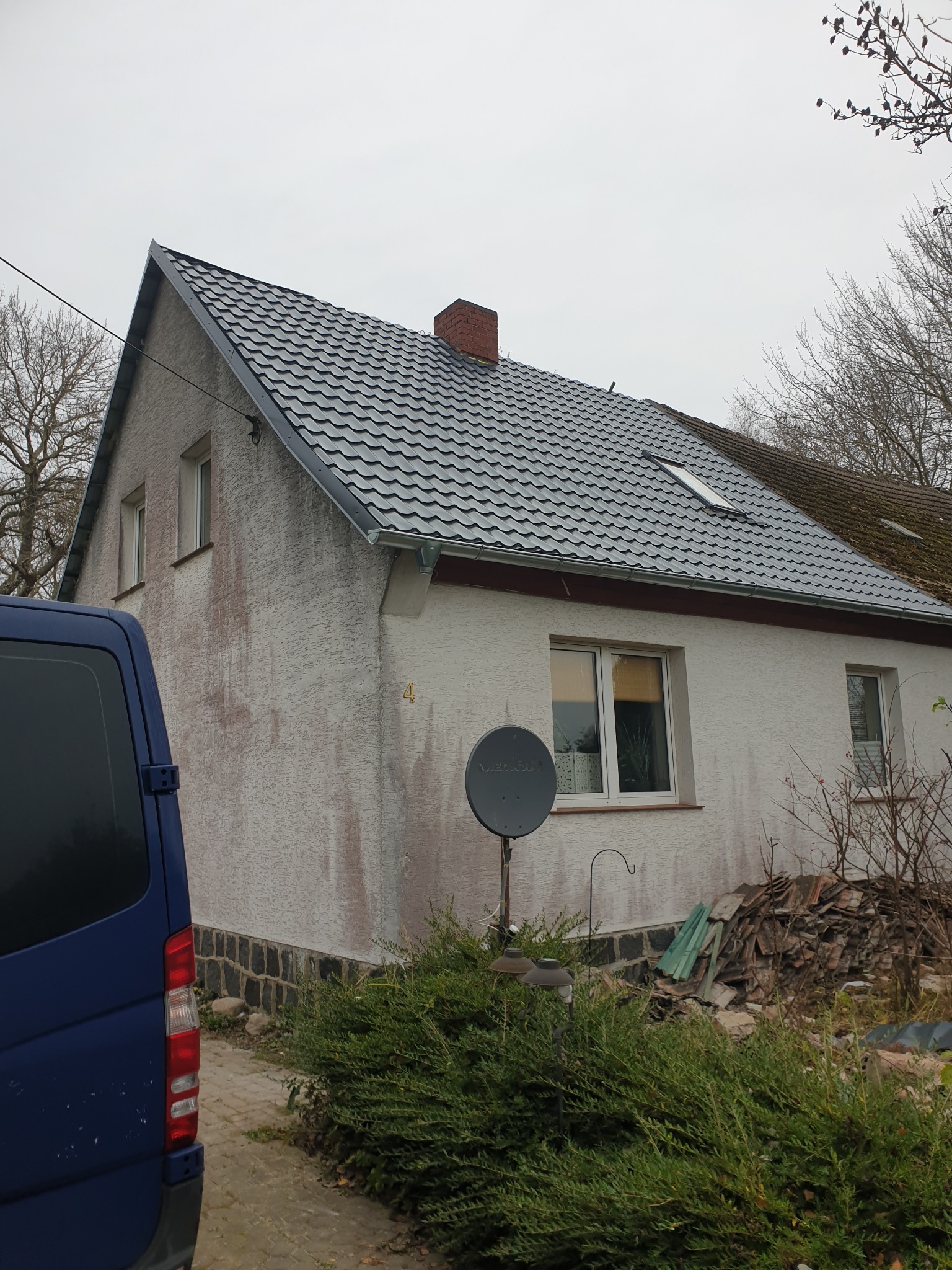 Dom jednorodzinny z częściowo nowym, szarym dachem z blachodachówki, widoczne okno dachowe i rynna, obok stara, zarośnięta mchem dachówka, elewacja budynku brudna, na pierwszym planie krzew...