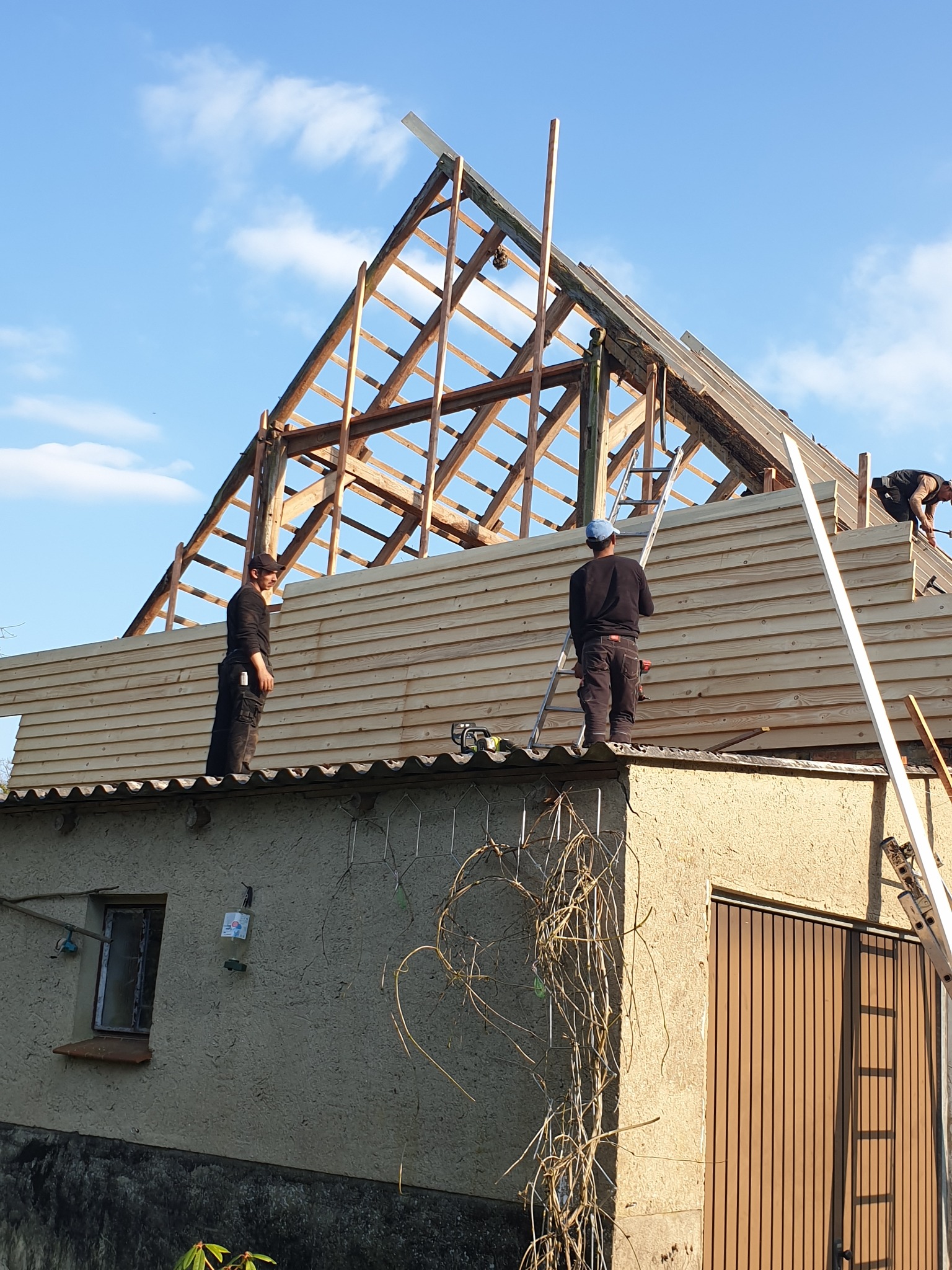 Ekipa dekarzy montuje drewnianą boazerię na dachu małego budynku z widoczną starą więźbą dachową i częściowo ułożonym pokryciem.