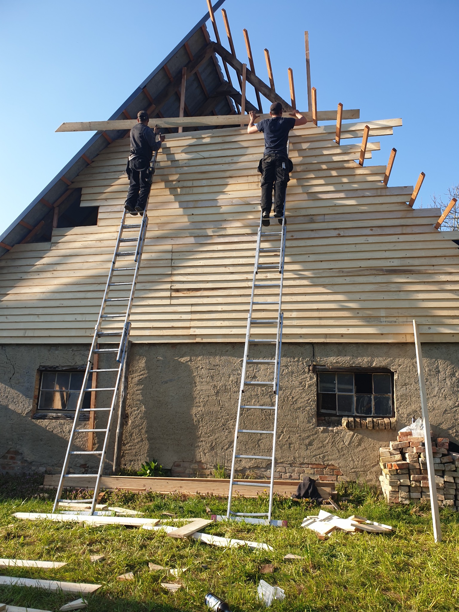 Dwóch pracowników na drabinach montuje drewnianą konstrukcję dachu na budynku z oknami, w tle błękitne niebo.