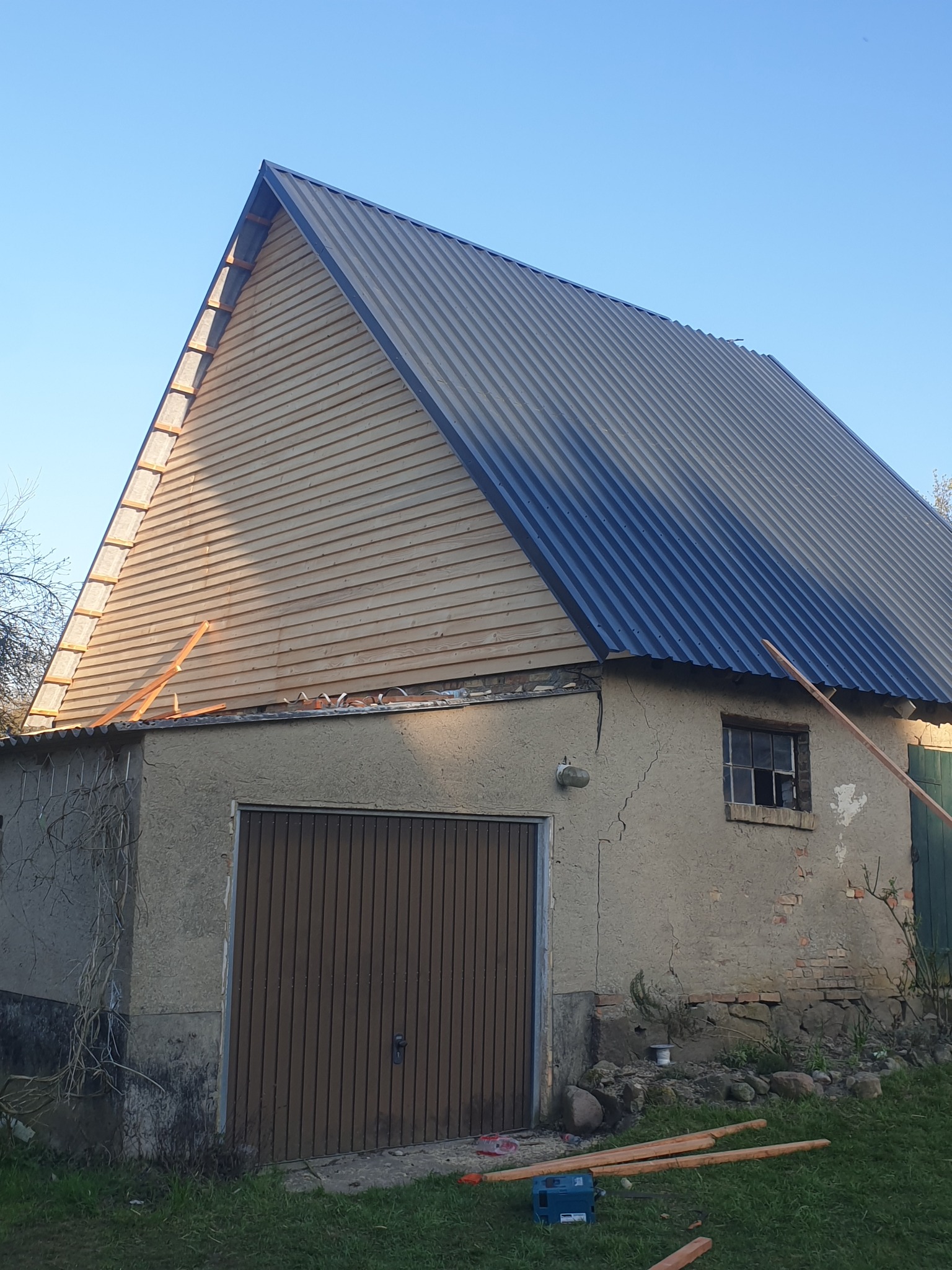 Budynek z częściowo wymienionym poszyciem dachowym z blachy trapezowej w kolorze grafitowym, z widoczną drewnianą konstrukcją szczytu i elementami łat na dachu. Stara, popękana elewacja budynku.