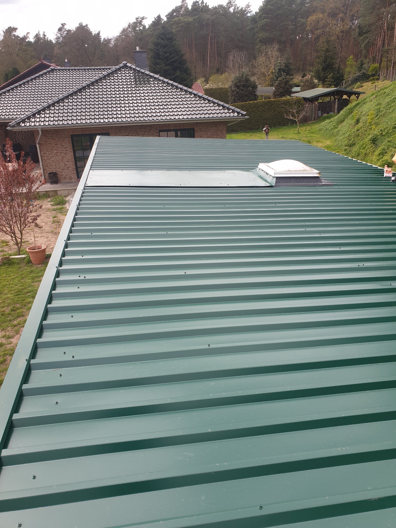 Zielony dach z blachy trapezowej z świetlikiem dachowym, widok z góry, w tle dom jednorodzinny i drzewa.