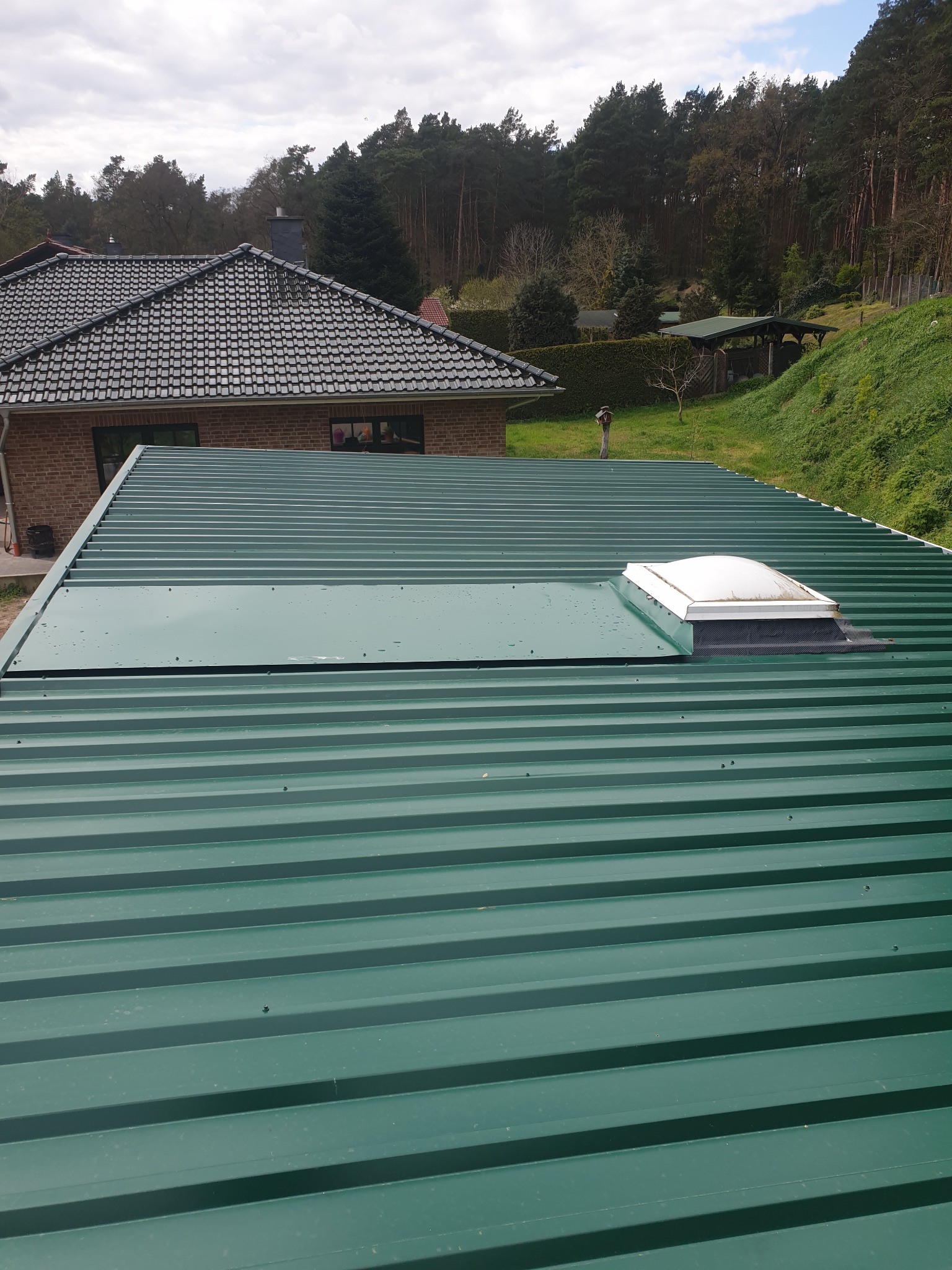 Nowy, zielony dach z blachy trapezowej z zamontowanym świetlikiem dachowym, widok z góry na tle domu i zieleni.