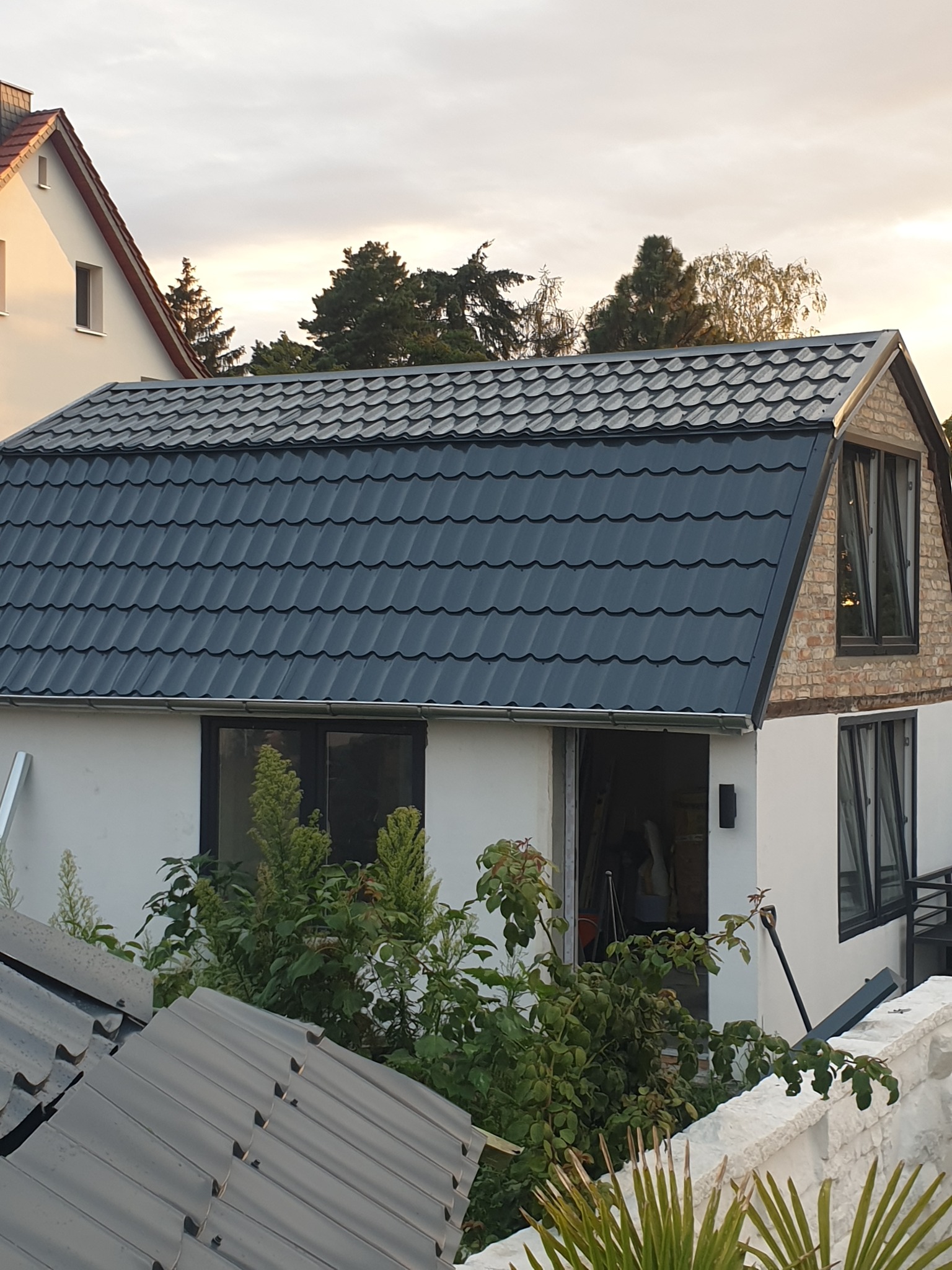 Nowy, grafitowy dach z blachodachówki na białym budynku z ceglaną nadbudówką poddasza i czarnymi oknami, widok z dołu na tle pochmurnego nieba.