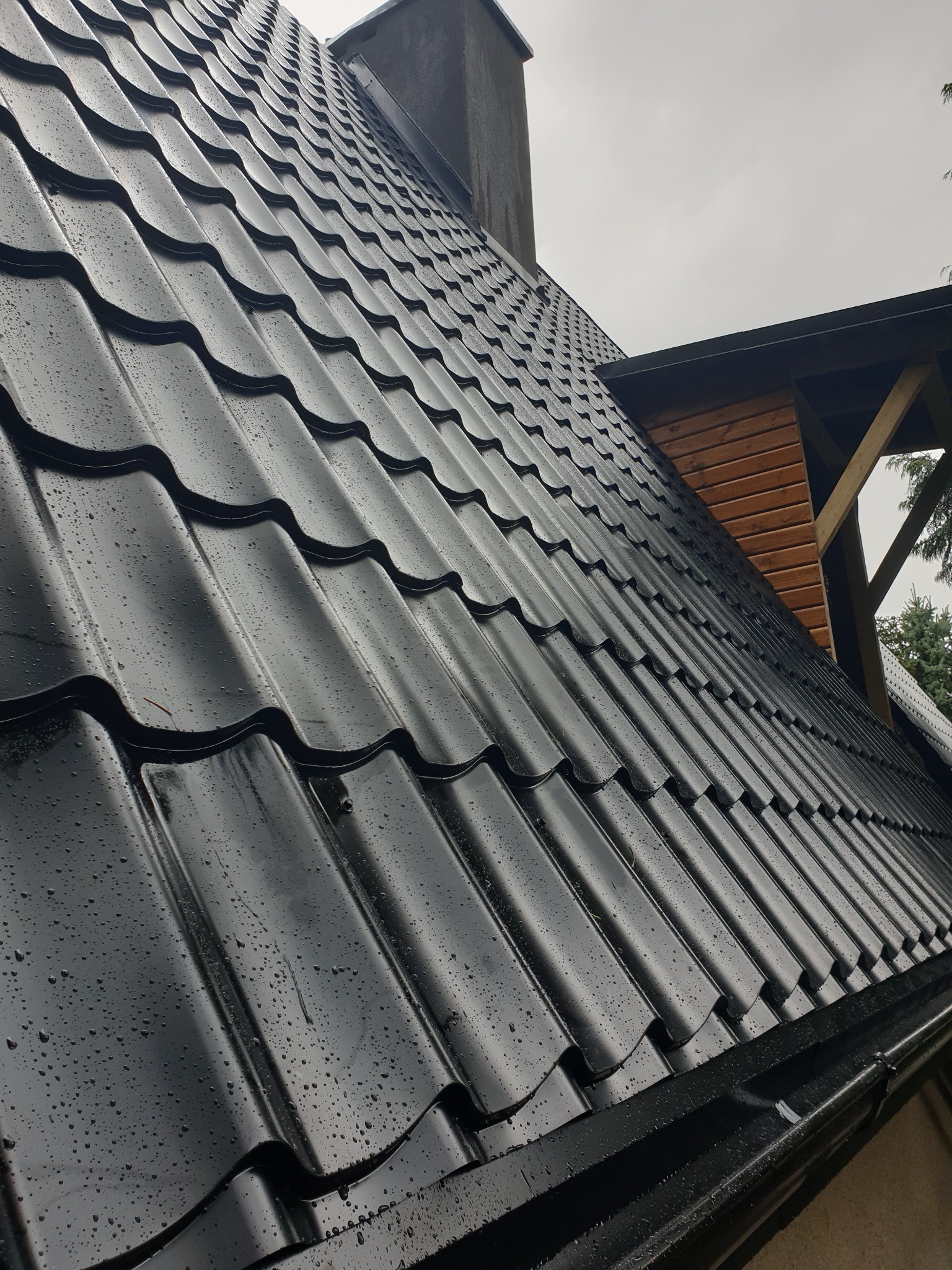 Nowy, czarny dach z blachodachówki, z widocznymi kroplami deszczu, zamontowany w Szczecinie, widok z dołu na fragment dachu i komina.