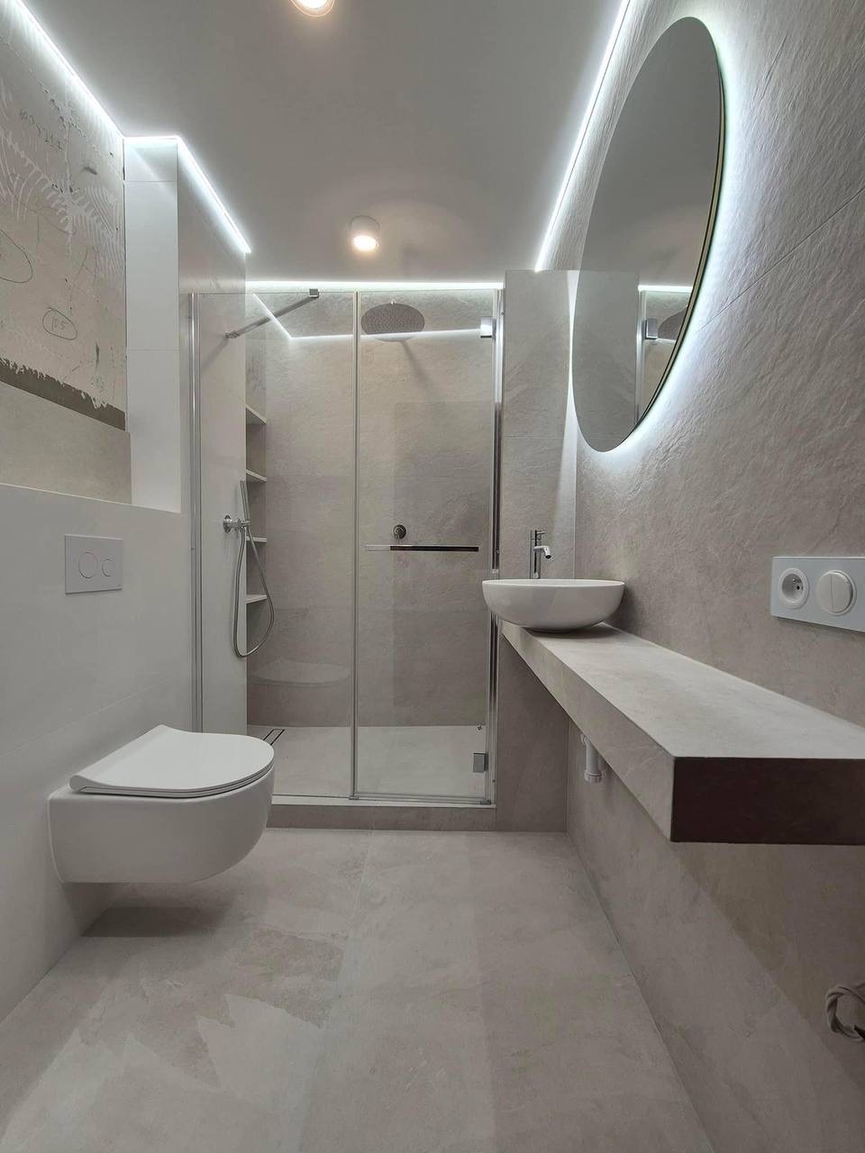 Nowoczesna łazienka w odcieniach szarości z podświetlanym lustrem, wiszącą toaletą i kabiną prysznicową typu walk-in, oświetlenie LED na suficie i ścianach.
