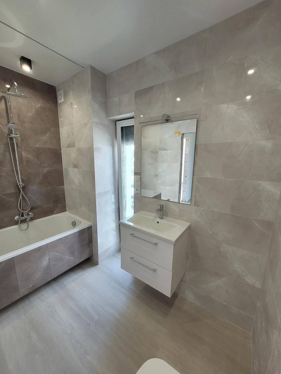 Jasna łazienka z marmurowymi płytkami, wisząca szafka z umywalką, bateria umywalkowa, wanna z obudową z płytek i deszczownica.
