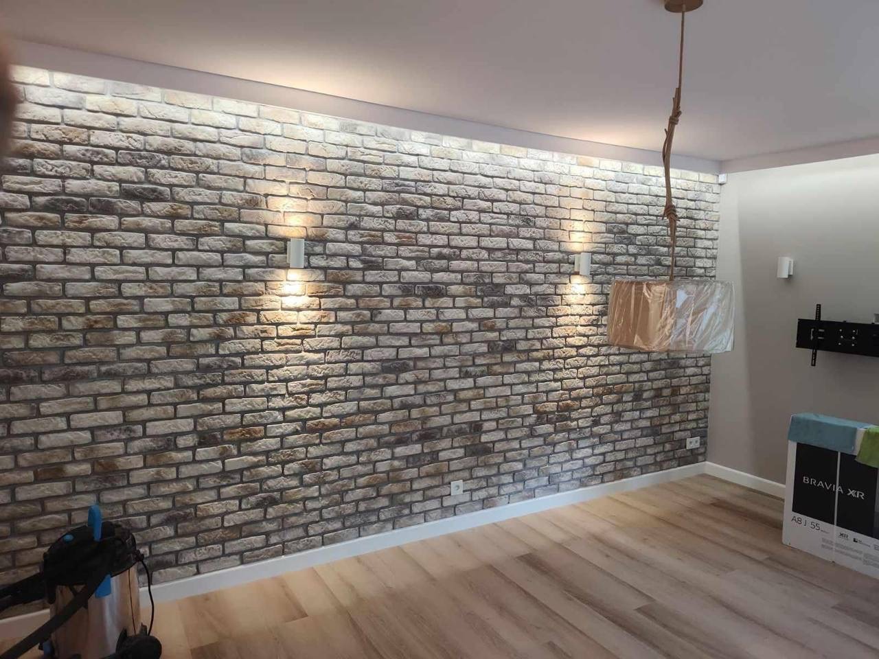 Ściana wykończona dekoracyjną cegłą z podświetleniem LED, drewniana podłoga, wisząca lampa z abażurem w folii, odkurzacz przemysłowy w rogu, karton po telewizorze Bravia XR