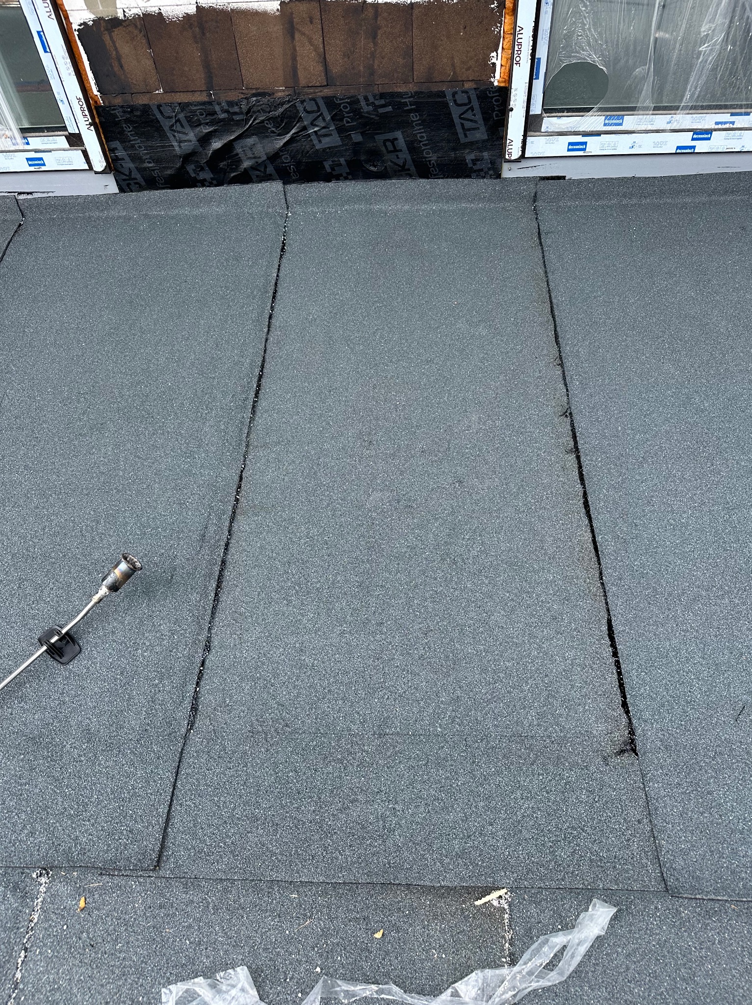 Widok z góry na fragment dachu pokrytego papą termozgrzewalną, z widocznymi łączeniami i palnikiem dekarskim w lewym dolnym rogu. Widoczne okno dachowe z obróbką blacharską.