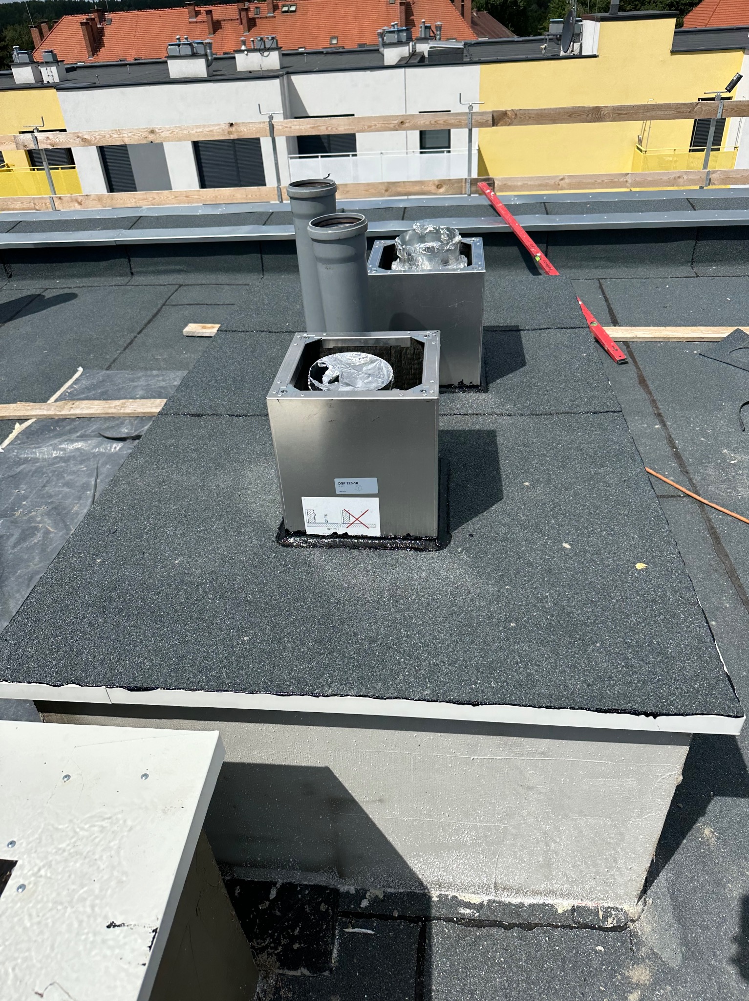 Montaż wentylacji na dachu pokrytym papą termozgrzewalną, widoczne dwa pionowe przewody wentylacyjne i dwa metalowe kominki wentylacyjne, jeden z folią aluminiową wewnątrz, poziomica w tle.