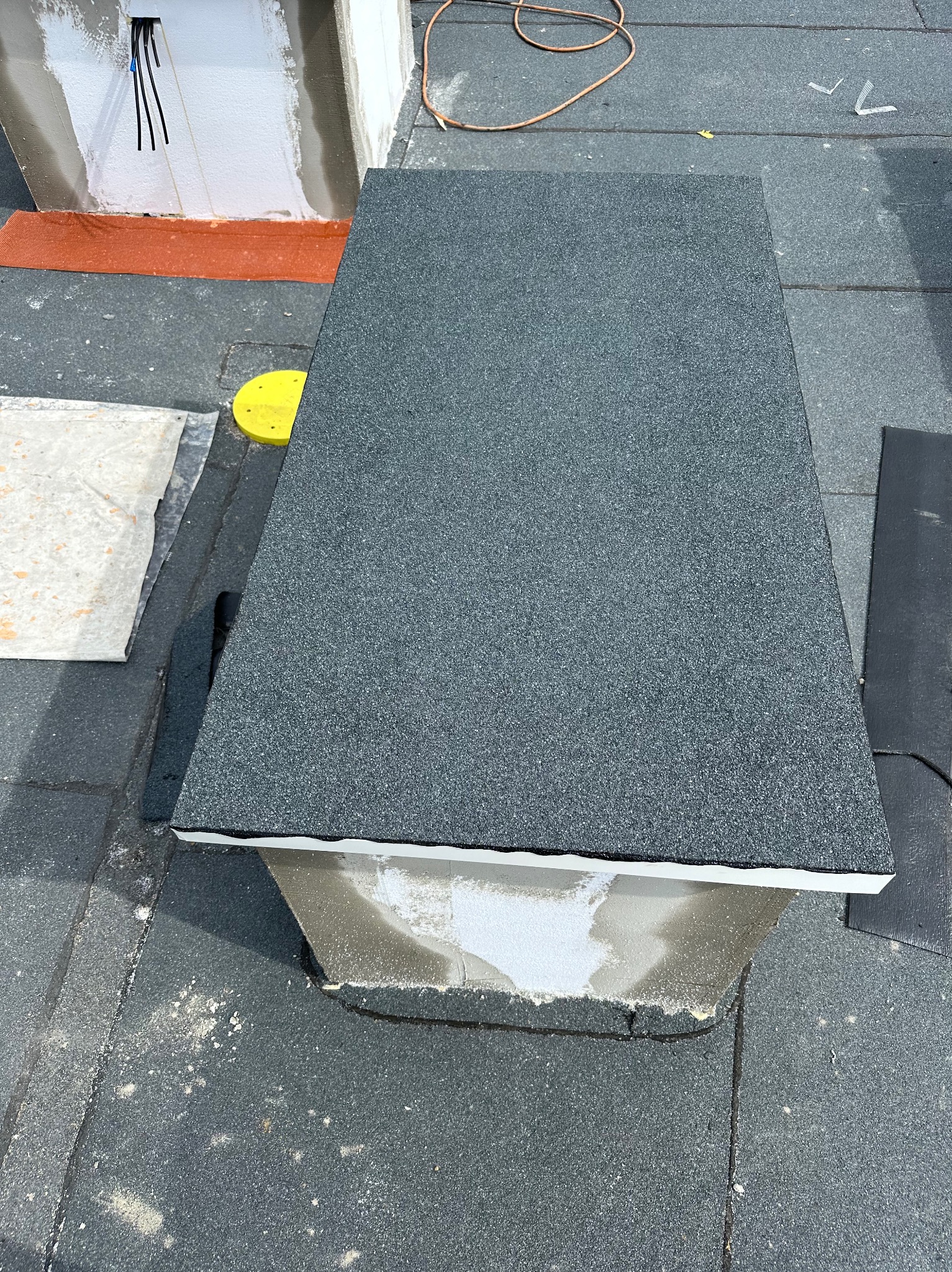 Szczegółowe ujęcie obróbki blacharskiej komina na dachu pokrytym papą, z widocznymi elementami uszczelniającymi i fragmentem instalacji elektrycznej.