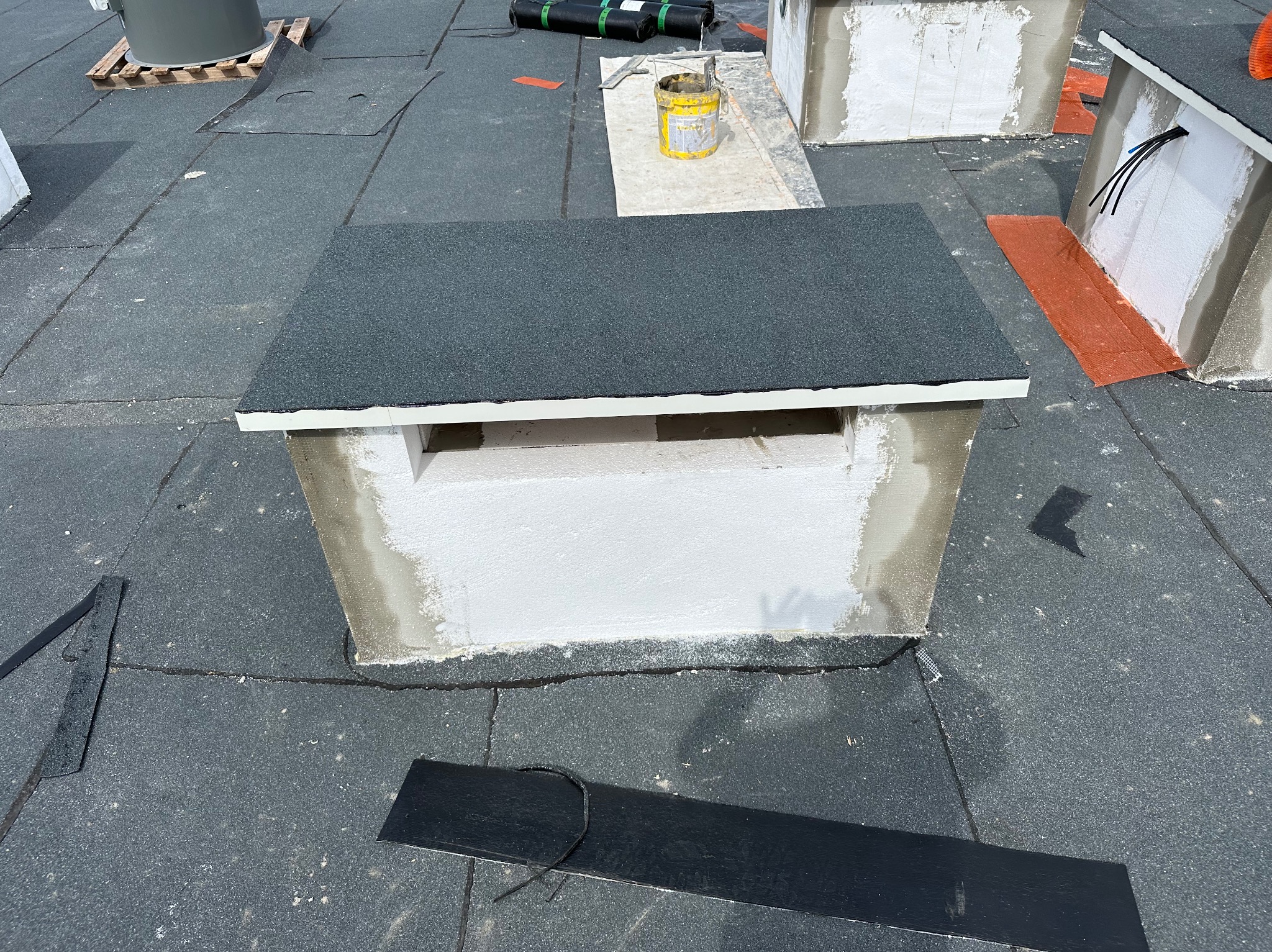 Wykończony komin wentylacyjny na płaskim dachu pokrytym papą, widoczne obróbki i uszczelnienia, w tle narzędzia i materiały budowlane.