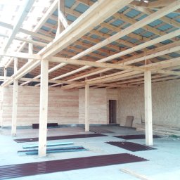 BIURO TRIVEN SP. Z O.O. - Wnętrze w trakcie budowy: drewniana konstrukcja szkieletowa domu z widocznymi elementami ścian z desek i płyt OSB, oraz przygotowane blachy dachowe na podłodze