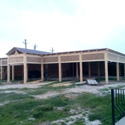 BIURO TRIVEN SP. Z O.O. - Konstrukcja szkieletowa z drewna w trakcie budowy, widoczne kratownice, słupy, częściowo zadaszona, z elementami wykończeniowymi, na zewnątrz budynku, w tle zaparkowany samochód.