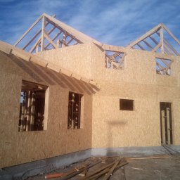 BIURO TRIVEN SP. Z O.O. - Surowy szkielet domu w trakcie budowy, ściany obite płytami OSB, widoczne drewniane elementy konstrukcyjne dachu i otworów okiennych, teren budowy z fragmentami drewna.