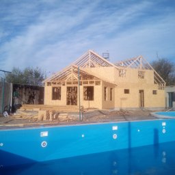 BIURO TRIVEN SP. Z O.O. - Wznoszenie szkieletu domu z płyty OSB na tle błękitnego, pustego basenu i pochmurnego nieba. Widoczne elementy konstrukcyjne dachu i ścian.