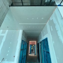 BIURO TRIVEN SP. Z O.O. - Korytarz w trakcie prac wykończeniowych, ściany i sufit pokryte płytami gipsowo-kartonowymi, zabezpieczone folią drzwi, widoczne instalacje pod sufitem.