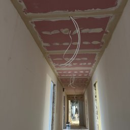 BIURO TRIVEN SP. Z O.O. - Korytarz w trakcie prac wykończeniowych, widoczny sufit z płyt kartonowo-gipsowych z widocznymi łączeniami i instalacją elektryczną, ściany przygotowane pod malowanie, w tle postać pracownika.