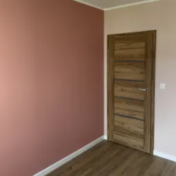 Pomieszczenie po malowaniu ścian na odcienie różu, z drewnianą podłogą i dopasowaną kolorystycznie stolarką drzwiową z poziomymi przeszkleniami.