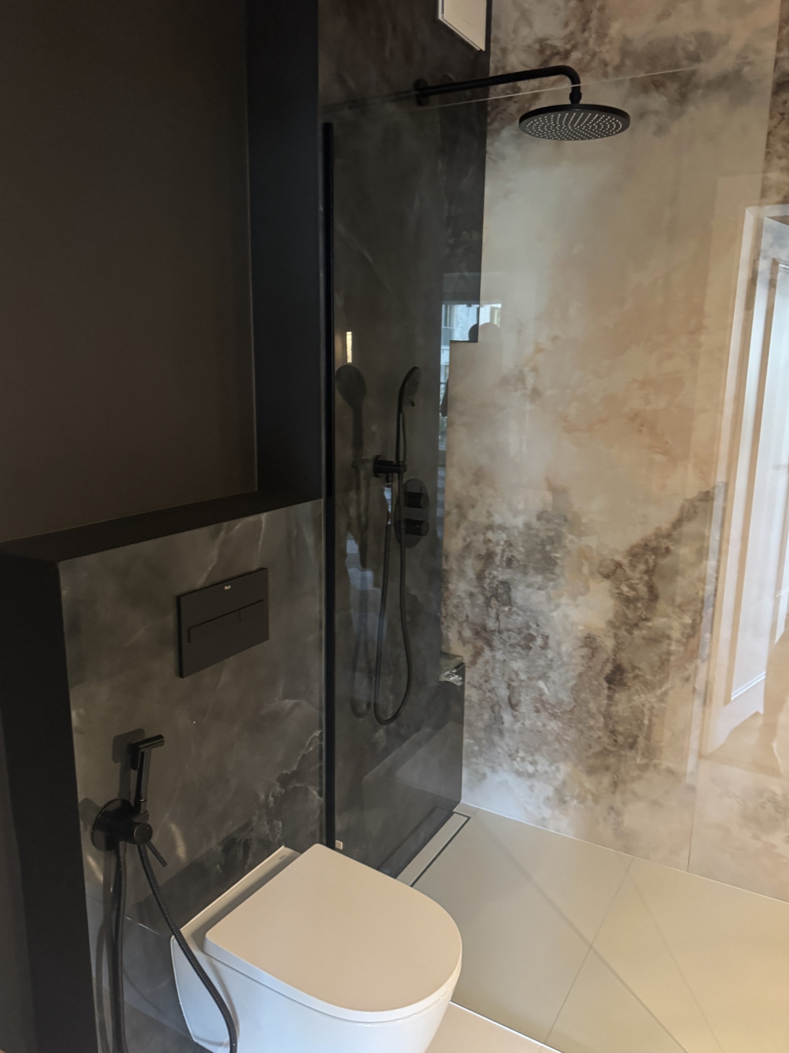 Nowoczesna łazienka z kabiną typu walk-in, wykończona płytami imitującymi marmur. Widoczna toaleta z bidetką i deszczownica w kolorze czarnym.