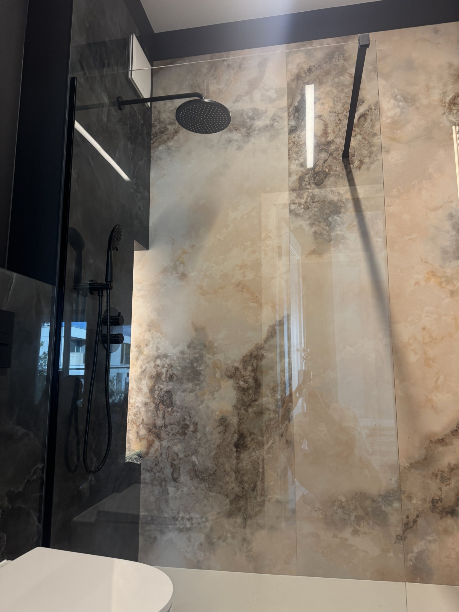 Nowoczesna kabina prysznicowa walk-in z czarną armaturą na tle marmurowych płytek w odcieniach beżu i szarości. Minimalistyczny design, widoczny fragment toalety.