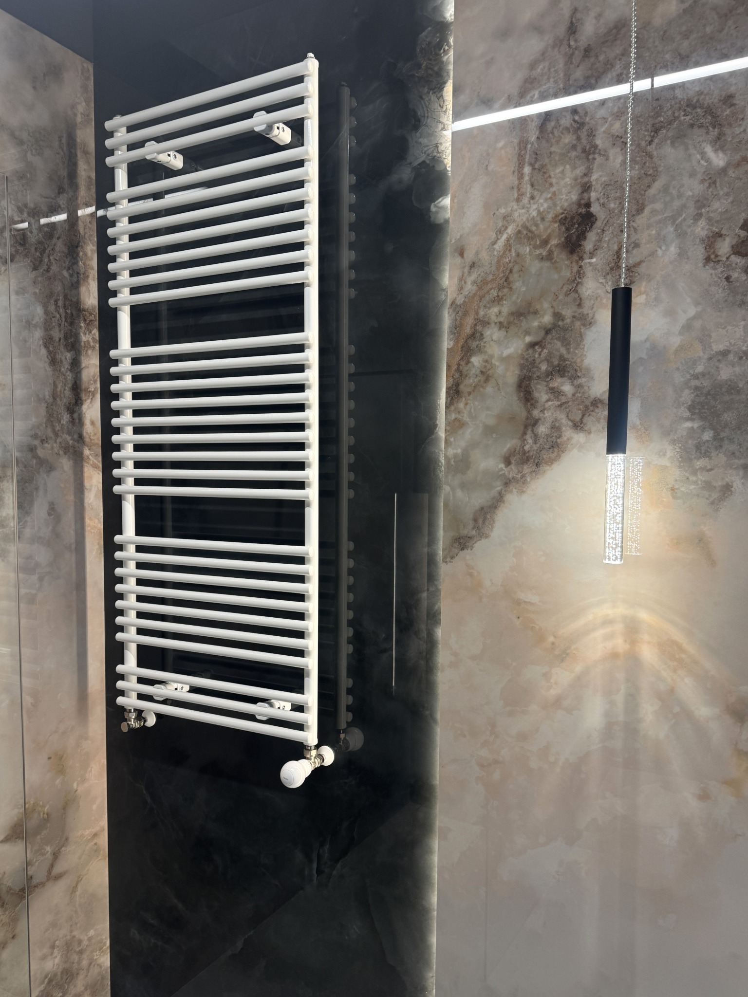 Biały grzejnik łazienkowy na tle marmurowej ściany i czarnego panelu, oświetlony wiszącą lampą. Nowoczesna aranżacja łazienki z dbałością o detale.