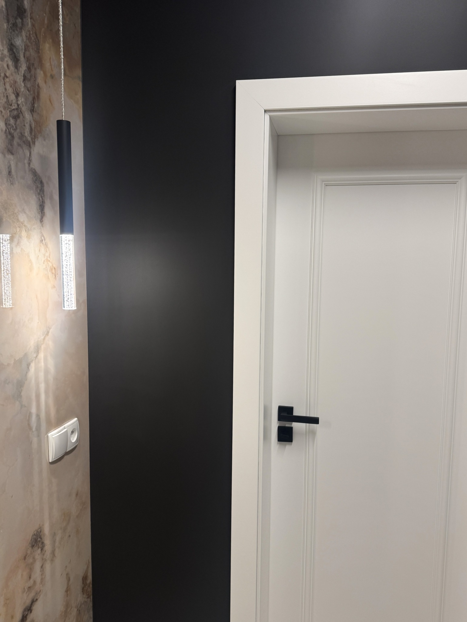 Nowoczesne wykończenie: białe drzwi z czarną klamką na tle ciemnej ściany, obok marmurowa ściana z podłużną lampą i gniazdkiem. Minimalistyczny design wnętrza.