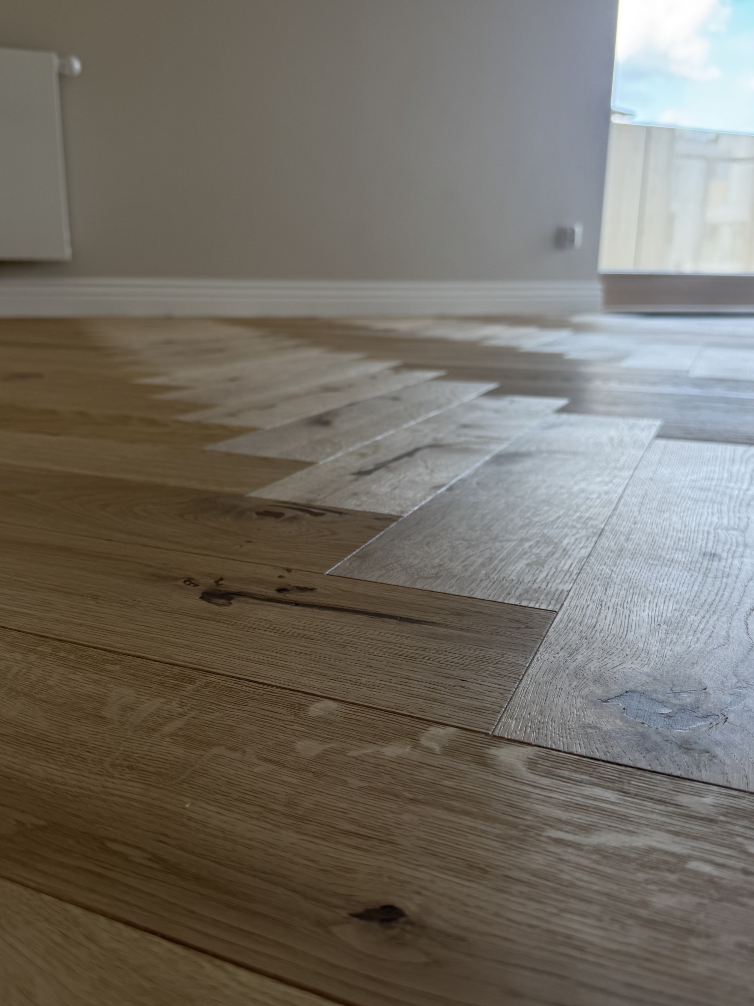 Parkiet ułożony w jodełkę francuską, z widocznymi słojami drewna i subtelnymi różnicami w odcieniach, tworzący elegancki wzór na podłodze w jasnym wnętrzu.