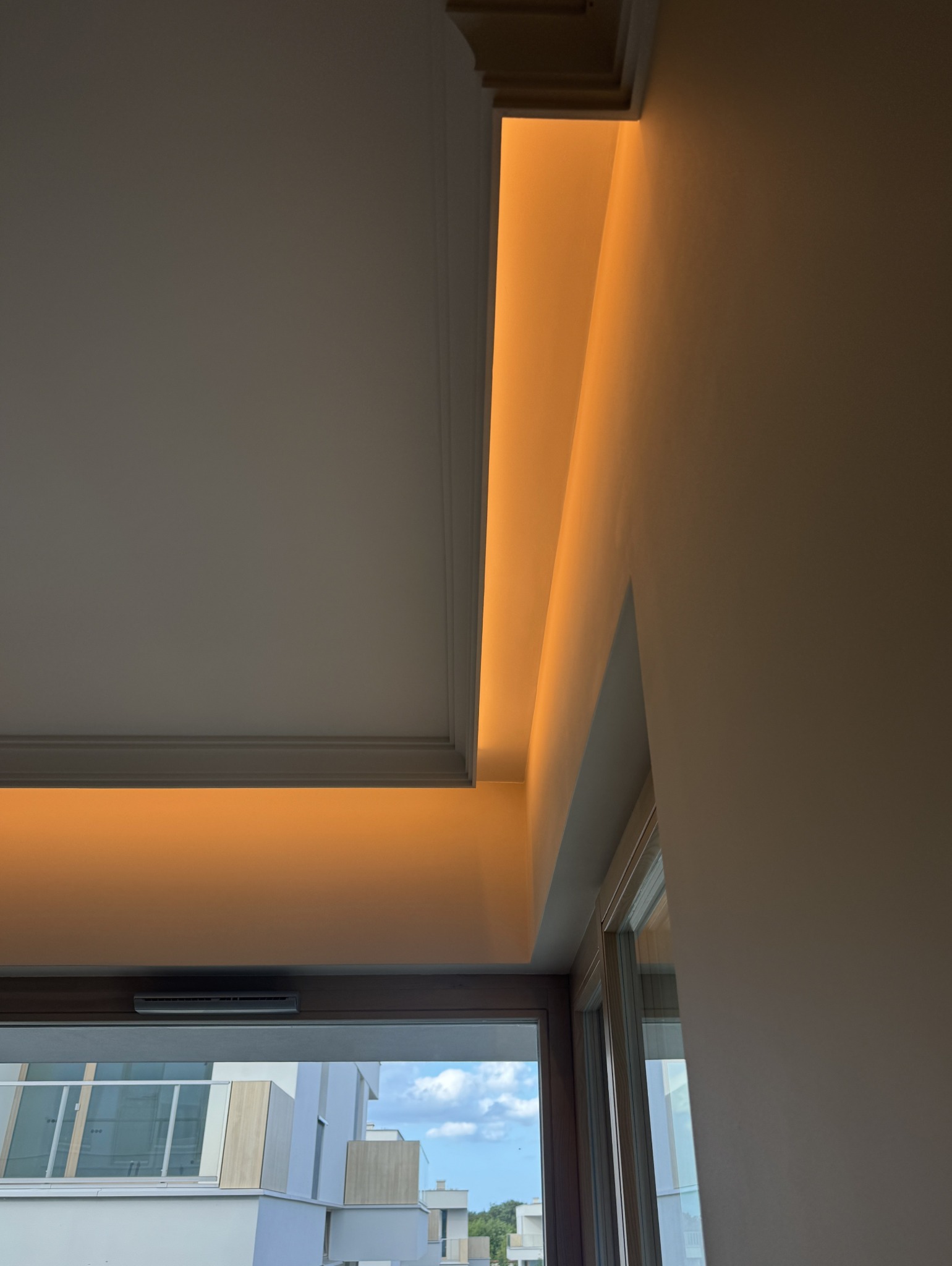 Nowoczesne oświetlenie sufitowe LED wnękowe, ciepła barwa światła, widok na okno i budynki w tle. Minimalistyczny design, eleganckie wykończenie wnętrza.