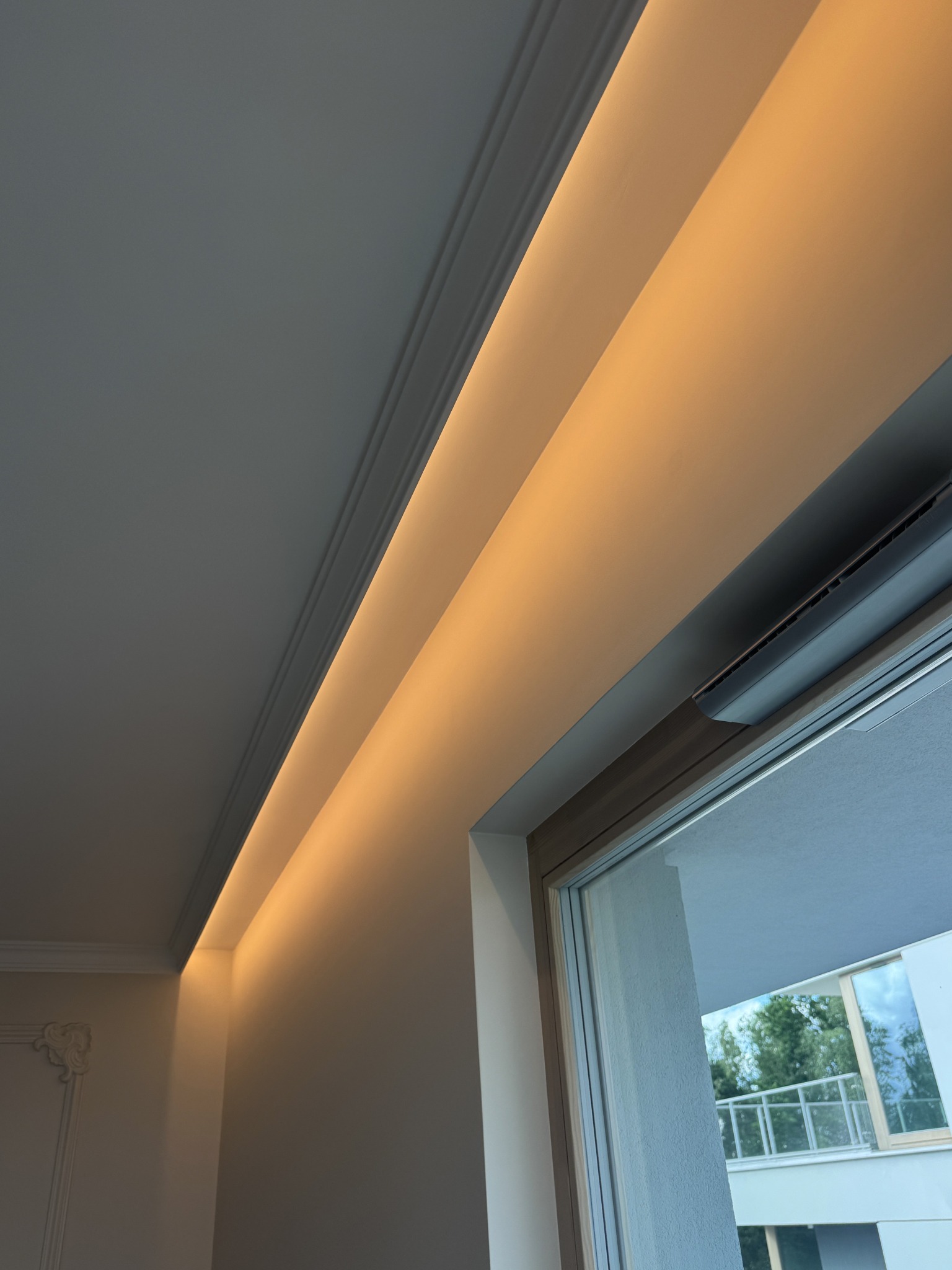 Oświetlenie LED w suficie podwieszanym z dekoracyjną listwą, ciepłe światło odbija się od białych ścian, widok na okno z klimatyzatorem.