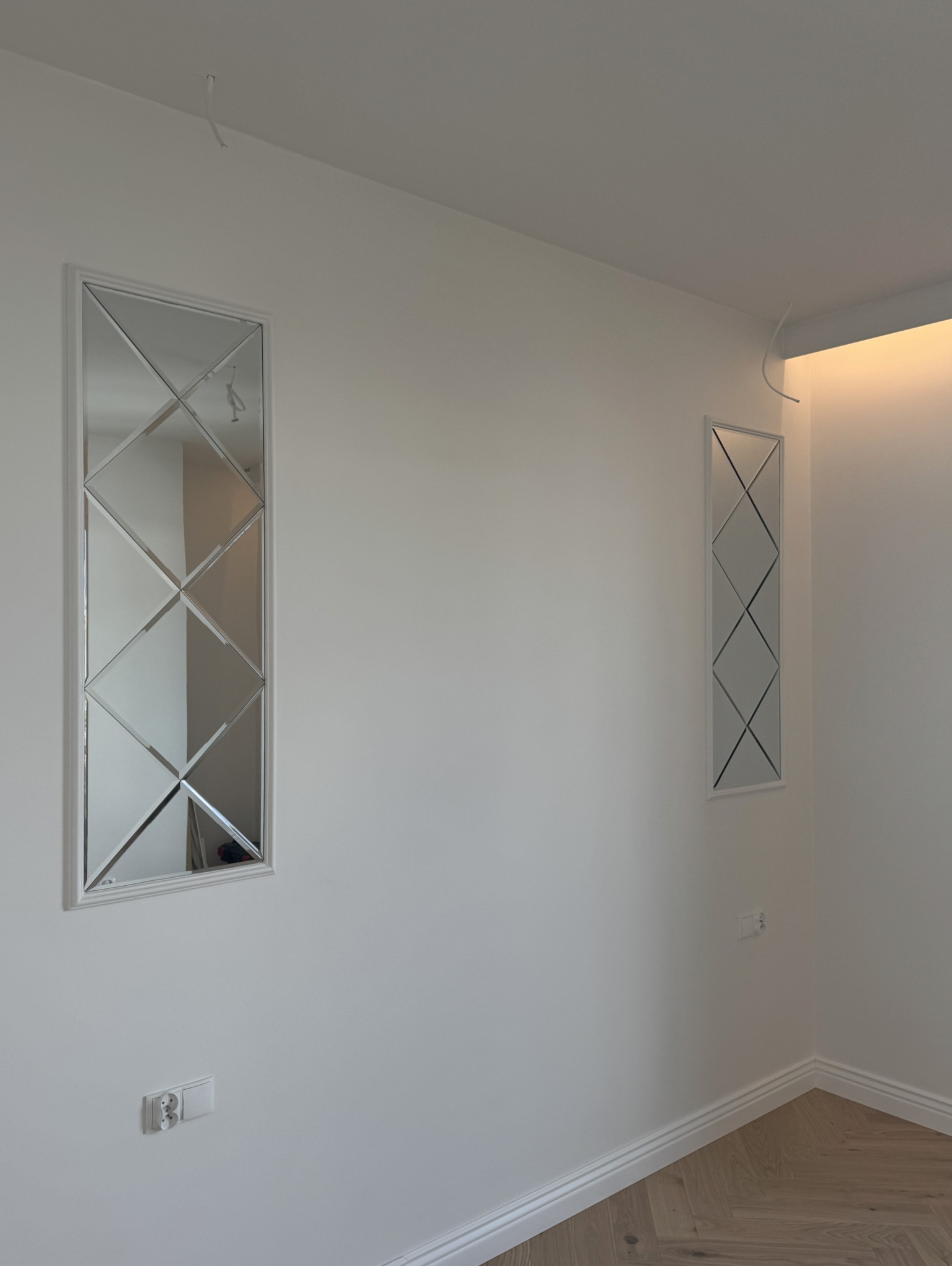 Dwa pionowe lustra w białych ramach z romboidalnymi podziałami na ścianie, parkiet na podłodze, widoczne gniazdka elektryczne, surowy kabel wystający z sufitu.