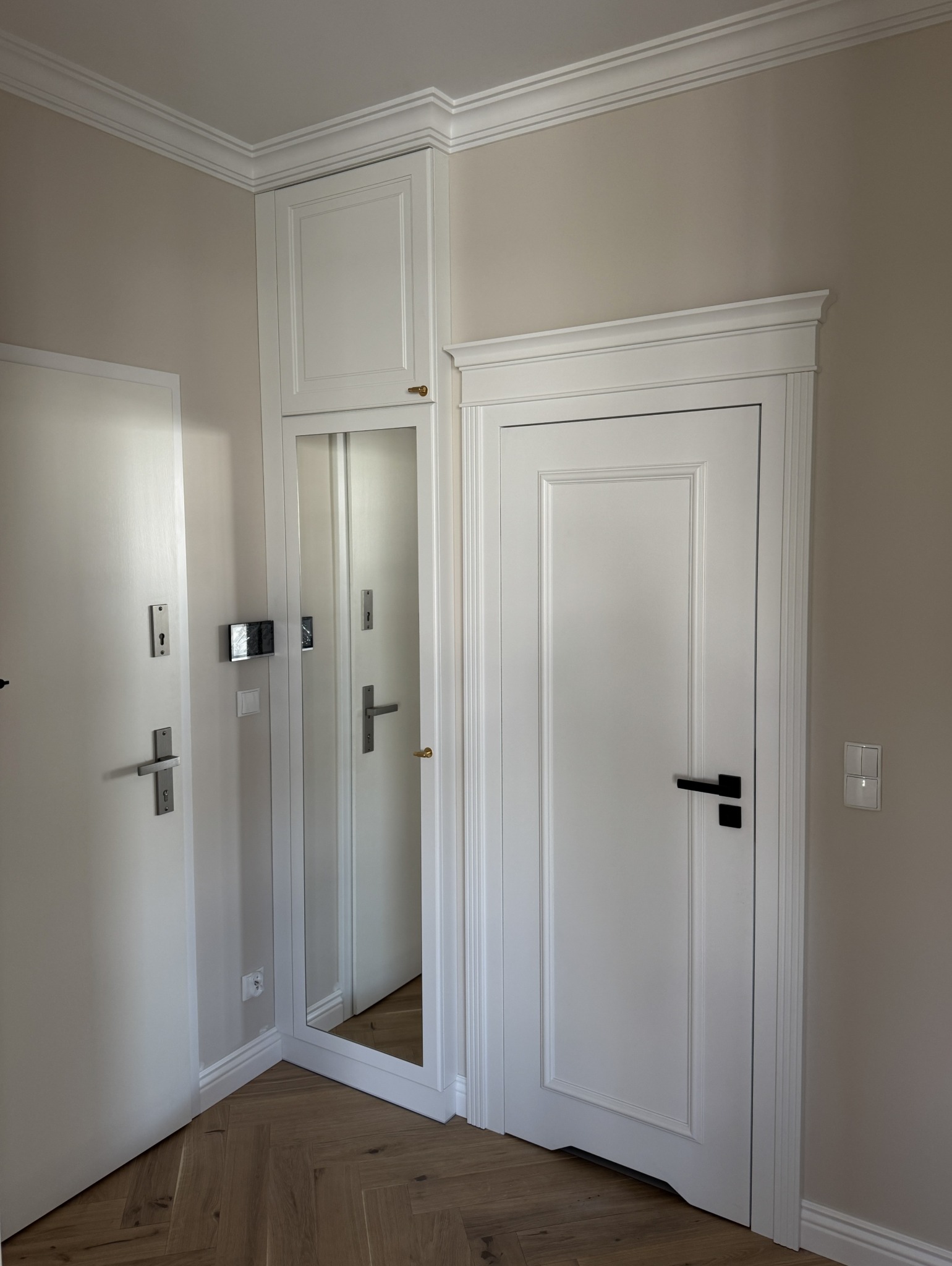 Białe drzwi wewnętrzne z ościeżnicą i szafa z lustrem w przedpokoju, wykończone listwami przysufitowymi. Parkiet ułożony w jodełkę. Styl klasyczny.
