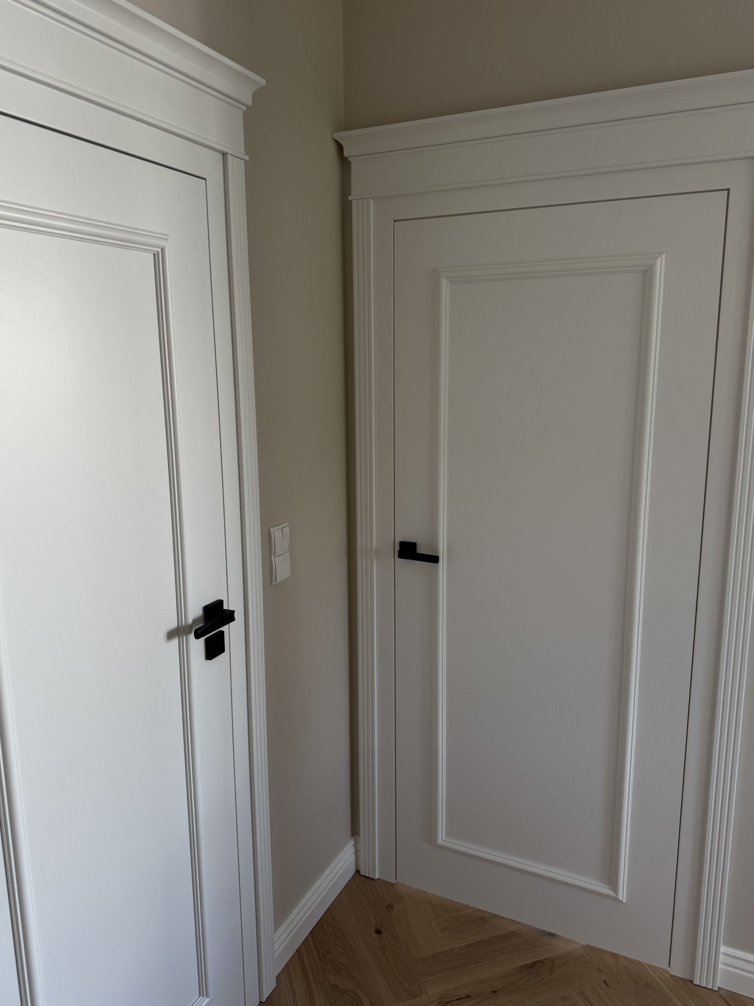 Dwa białe, klasyczne drzwi z czarnymi klamkami, wykończone listwami przypodłogowymi i ozdobnymi ramami. Parkiet w jodełkę na podłodze.