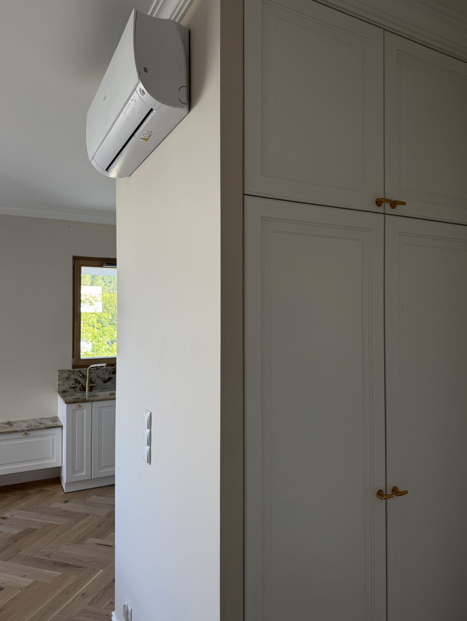 Klimatyzator na ścianie w Gdańsku, obok zabudowy meblowej z frezowanymi frontami i złotymi uchwytami. W tle widoczna podłoga w jodełkę i fragment okna.