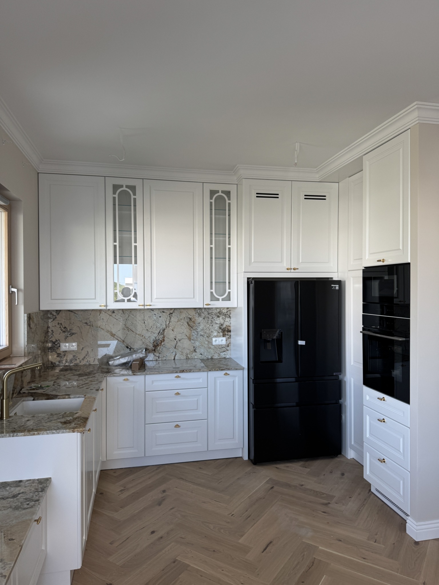 Elegancka, jasna kuchnia z marmurowym blatem i czarną lodówką. Parkiet w jodełkę. Szafki z witrynami i złotymi uchwytami. Nowoczesny design z klasycznymi elementami.