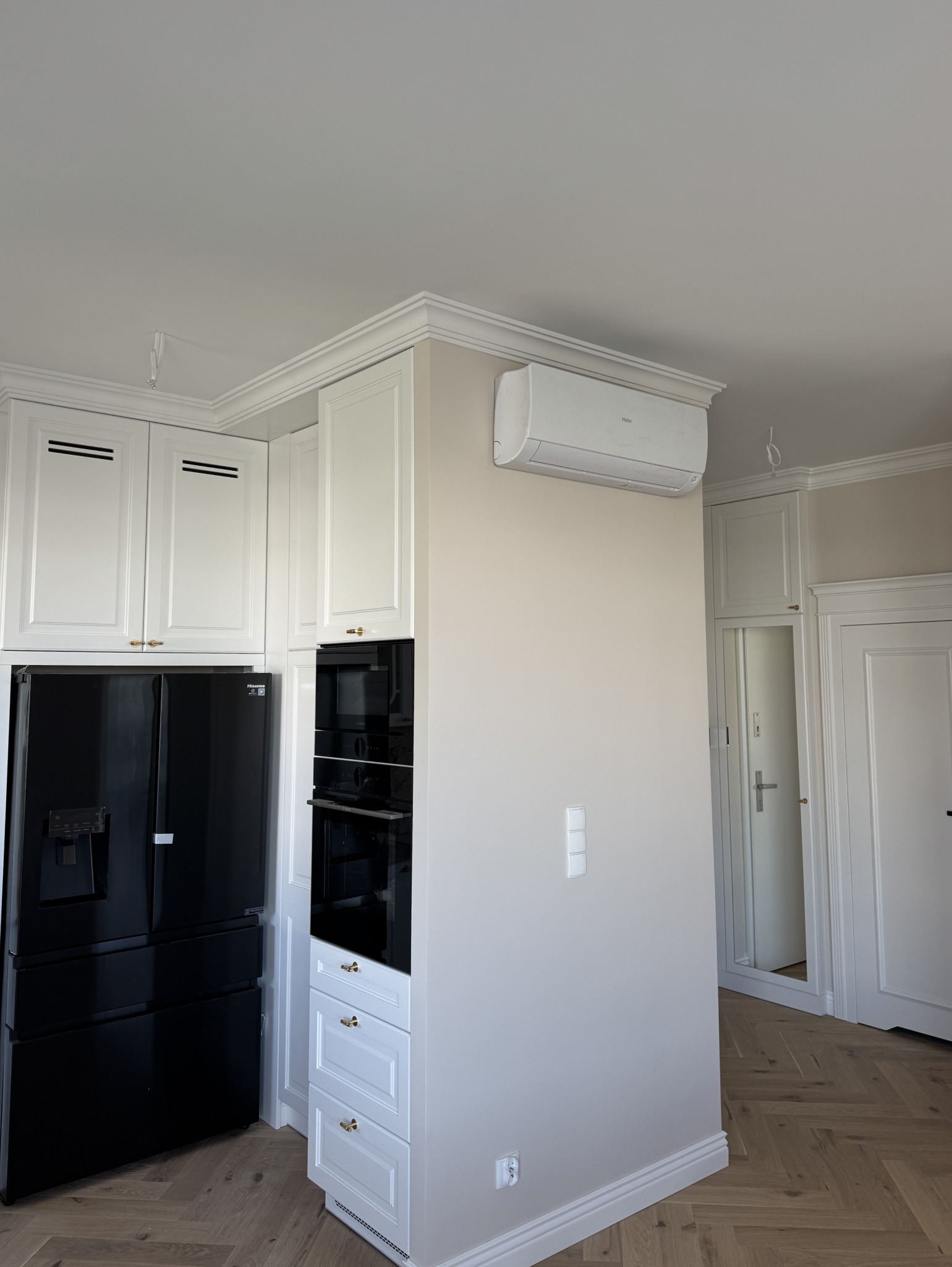 Nowoczesna kuchnia z białymi szafkami w stylu klasycznym, czarną lodówką i piekarnikiem, parkietem w jodełkę i klimatyzacją. Widok na drzwi z lustrem w tle.