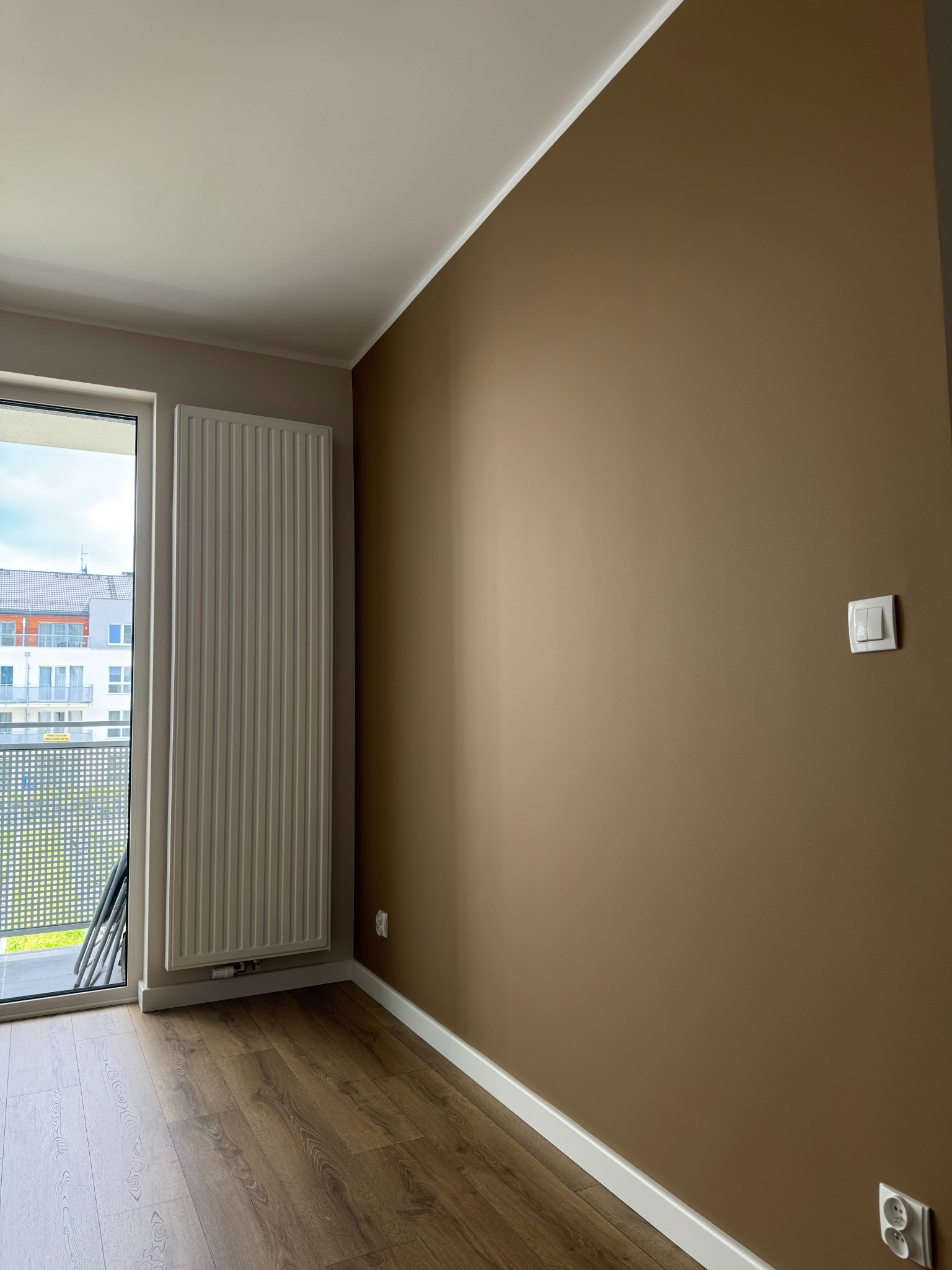 Pokój po malowaniu ścian na brązowo, z drewnianą podłogą i białymi listwami przypodłogowymi. Widoczny grzejnik i okno balkonowe z widokiem na inne budynki.
