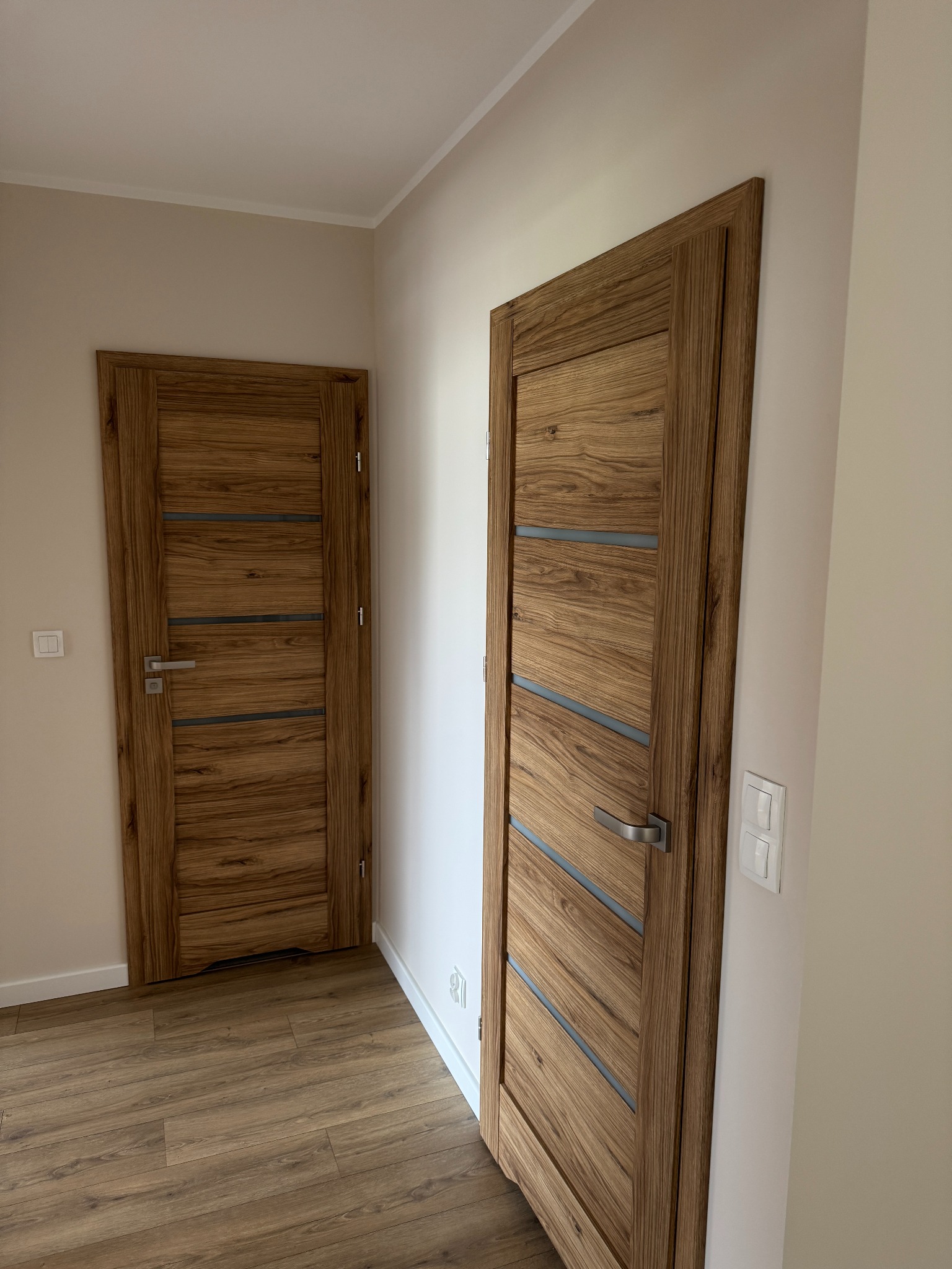Dwa drewniane drzwi wewnętrzne z poziomymi, szarymi wstawkami i srebrnymi klamkami, zamontowane w jasnym korytarzu z panelami podłogowymi.